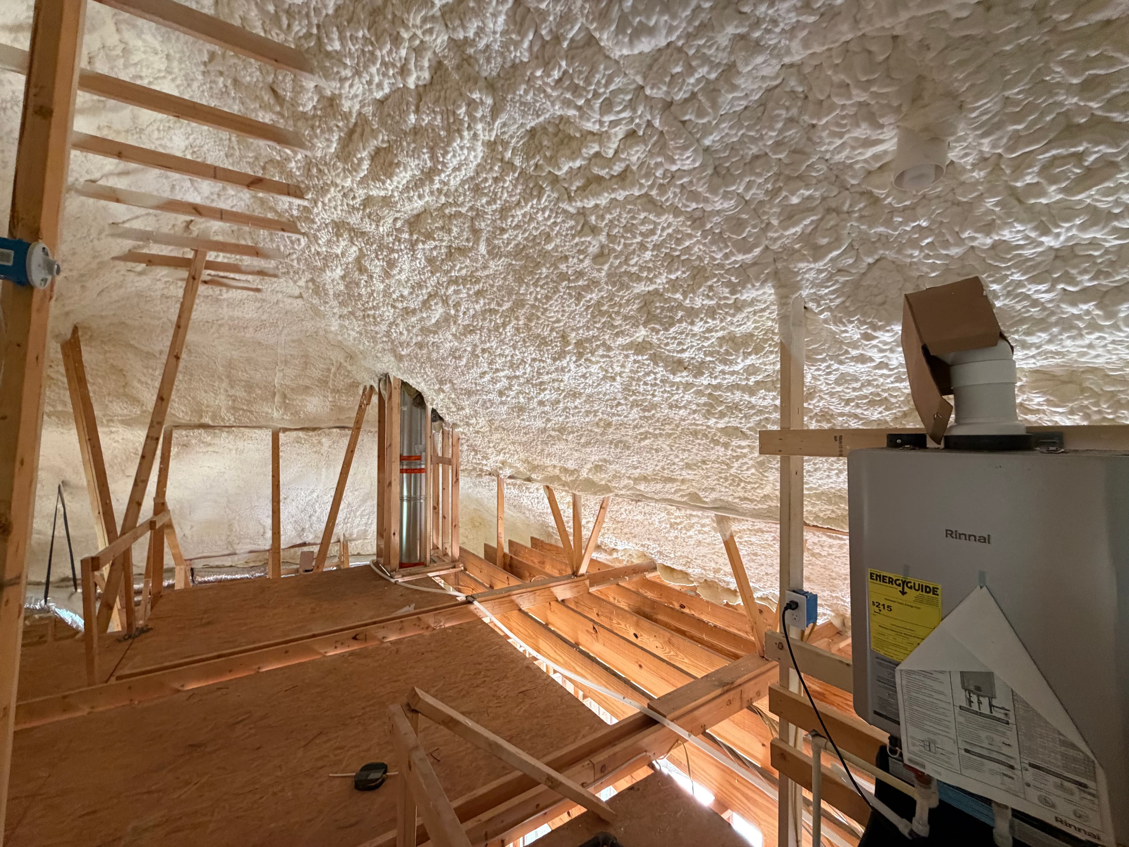 White spray foam insulation covers the ceiling and walls of a wooden framed attic.