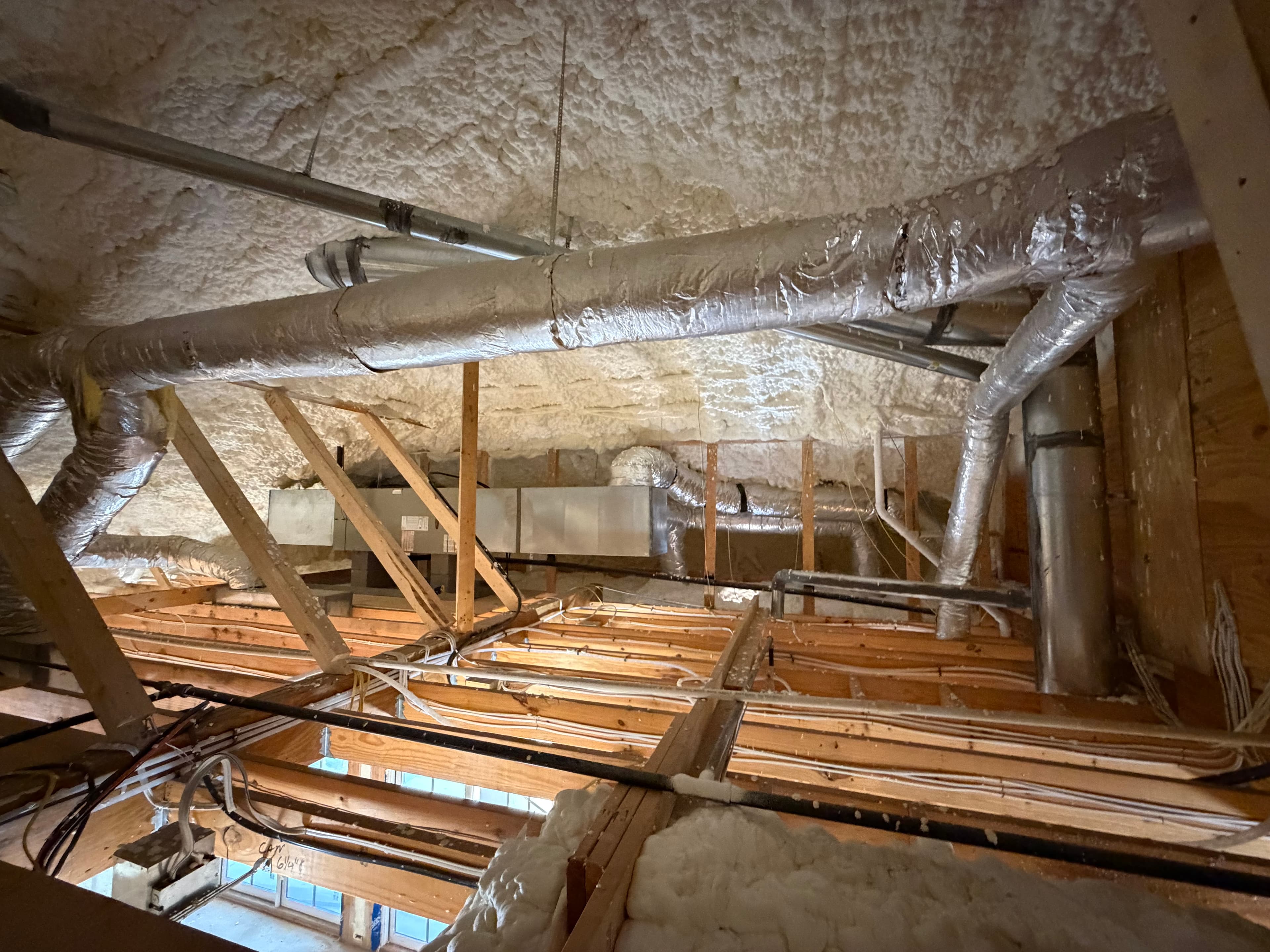 Attic interior featuring white spray foam insulation, wooden framing, and insulated silver HVAC ventilation ducts.