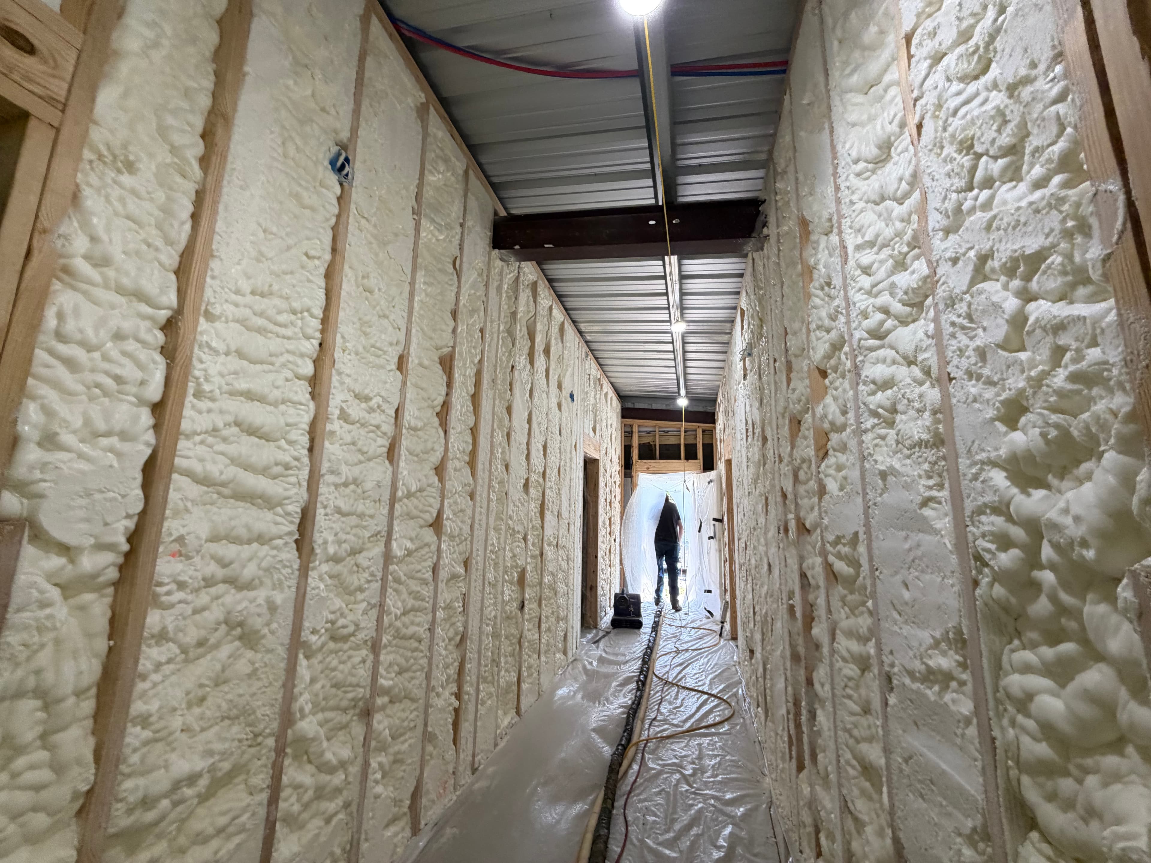 White spray foam insulation applied between wooden wall studs in a narrow hallway under construction.