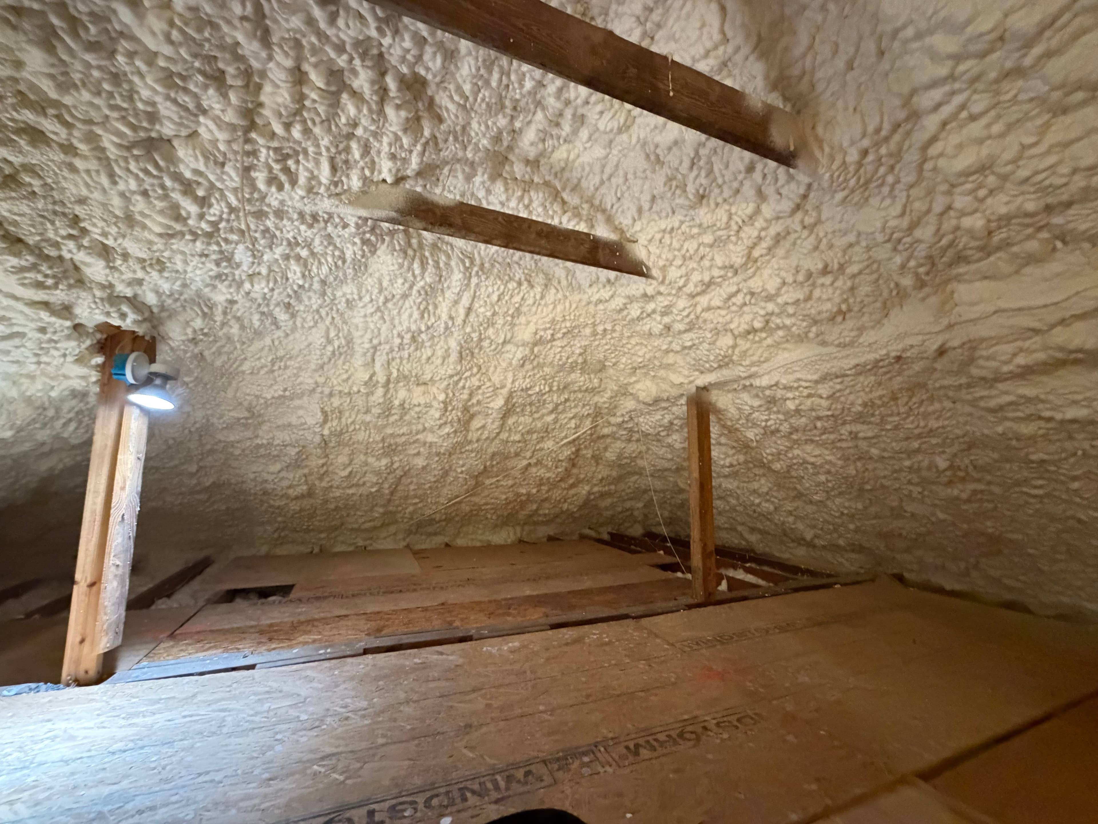 Thick white spray foam insulation covers the ceiling and walls of a wooden attic interior.