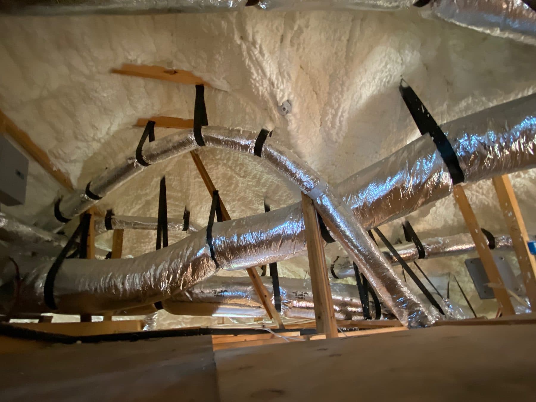 Flexible silver air ducts hanging in an attic with white spray foam insulation.