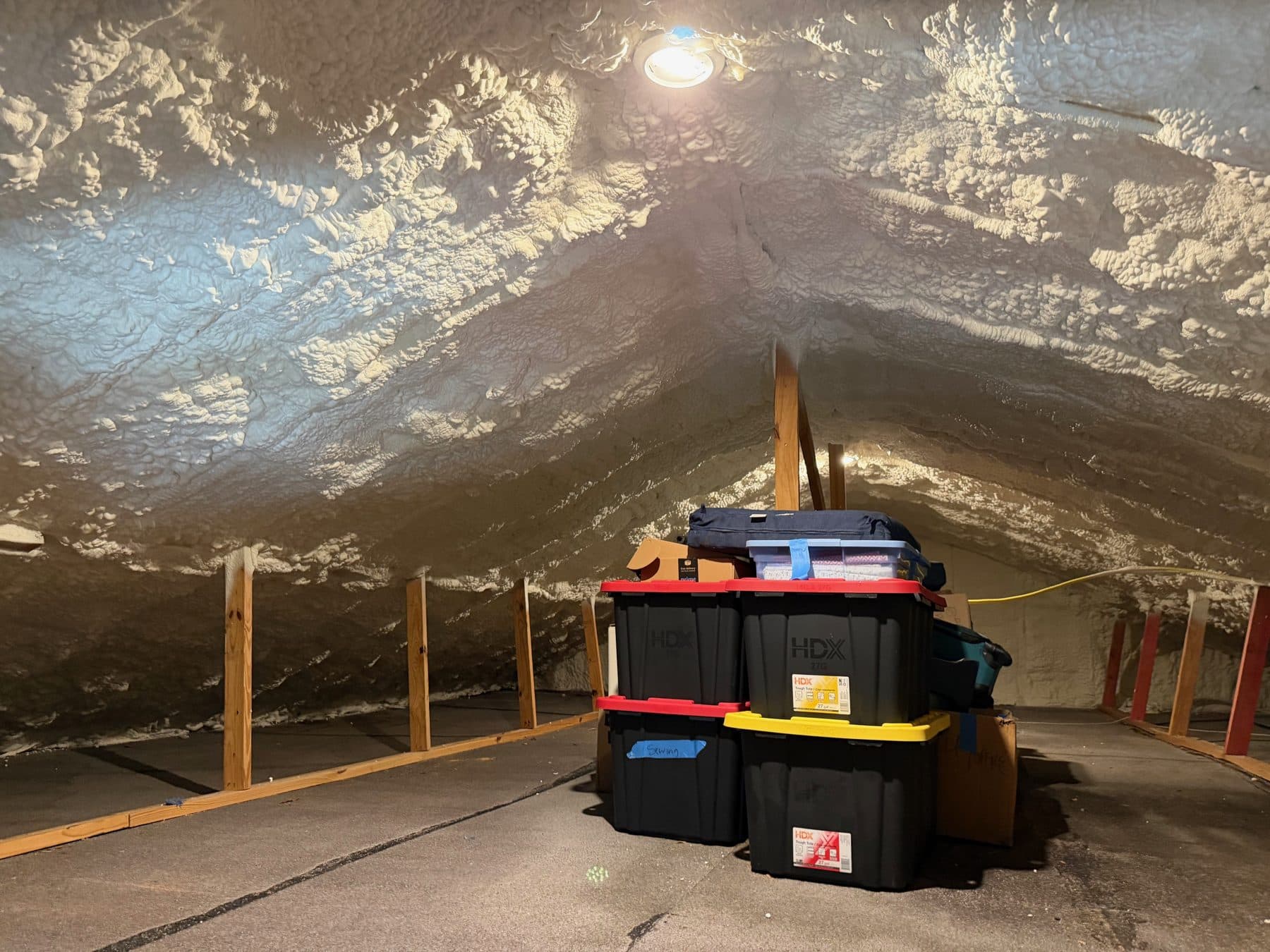 Attic with white spray foam insulation on the ceiling and walls, featuring stacked storage bins.