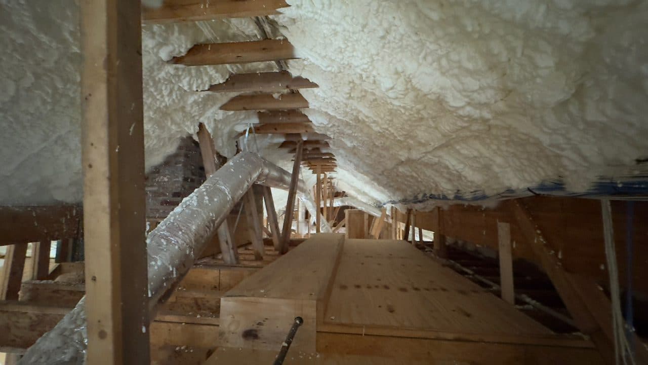 White spray foam insulation applied to wooden rafters in an attic with a silver duct.