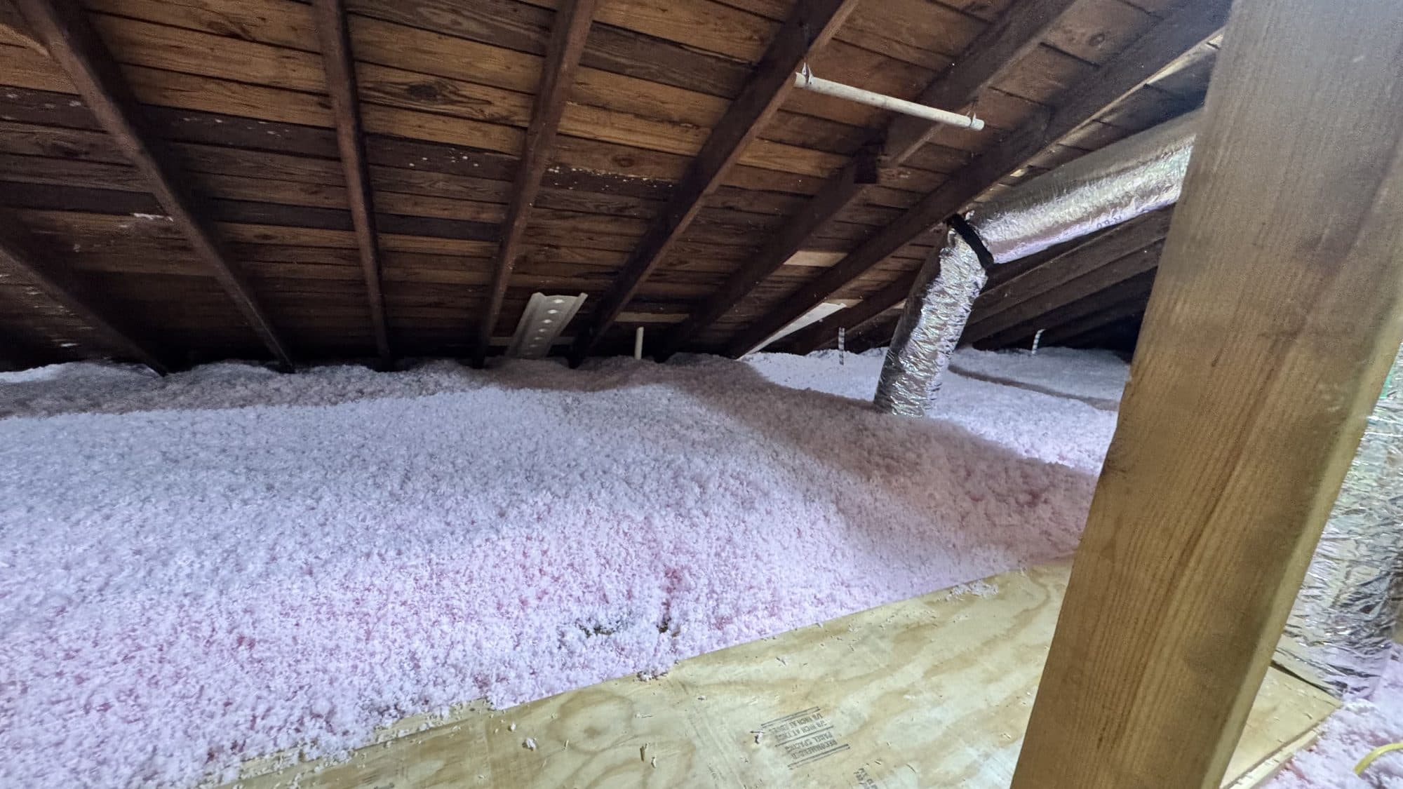 Pink blown-in insulation covers an attic floor beneath wooden rafters and a silver flexible duct.
