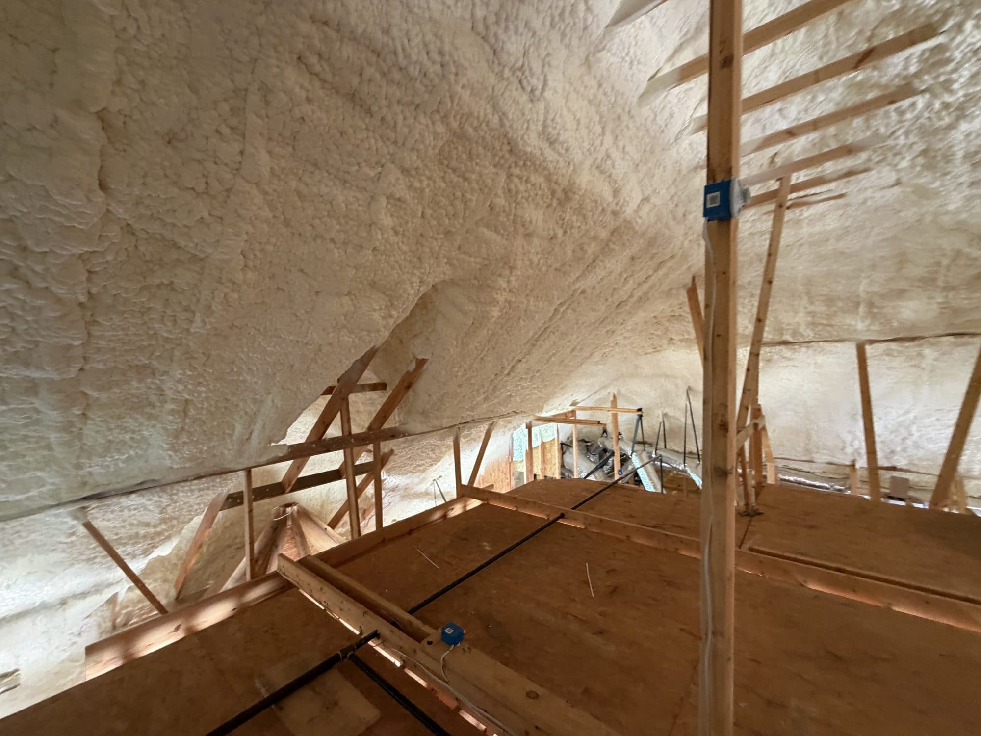 Attic interior with thick spray foam insulation on the ceiling and exposed wooden support beams.