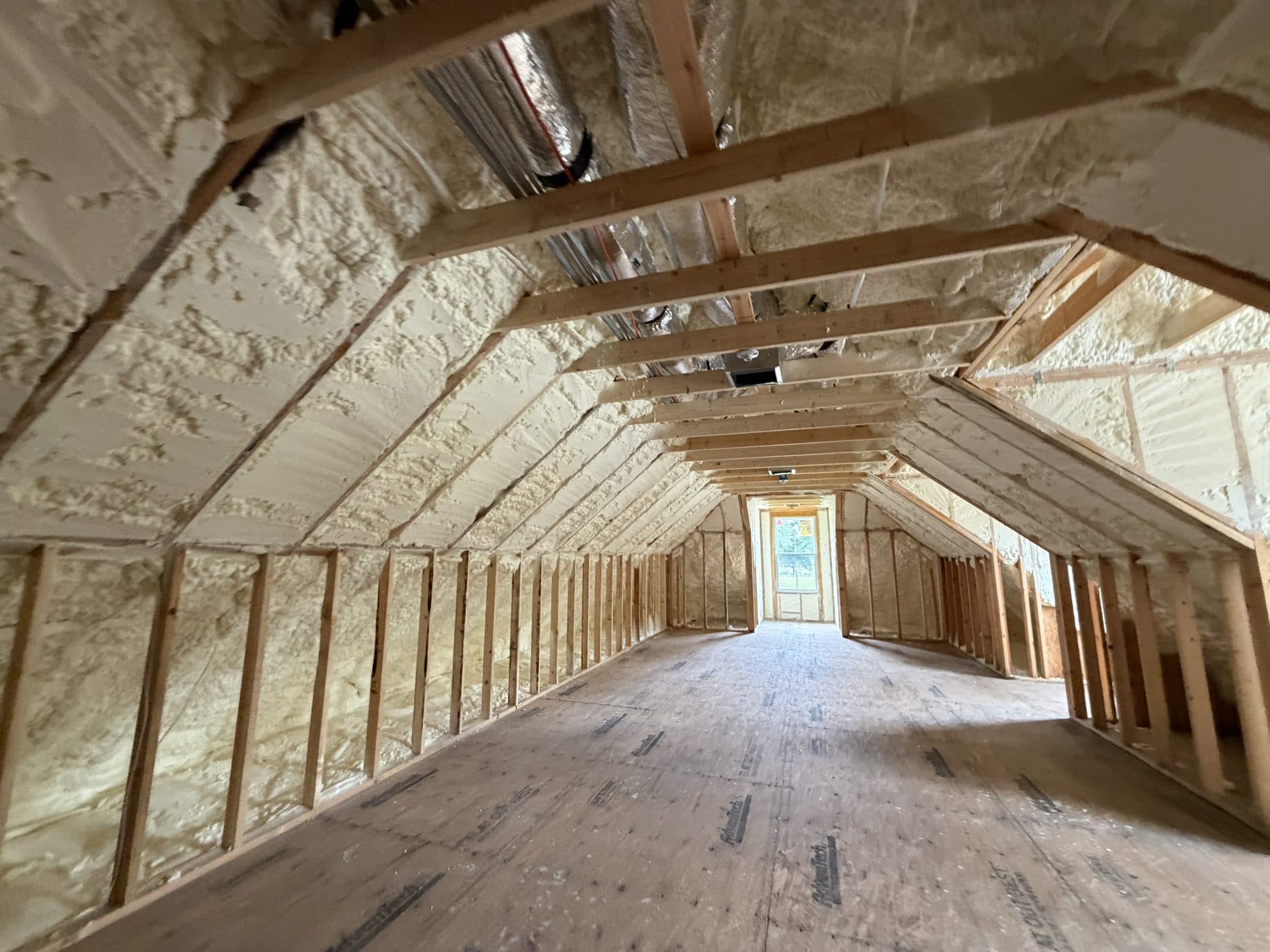 Unfinished attic with wooden framing, subflooring, and spray foam insulation on the walls and ceiling.