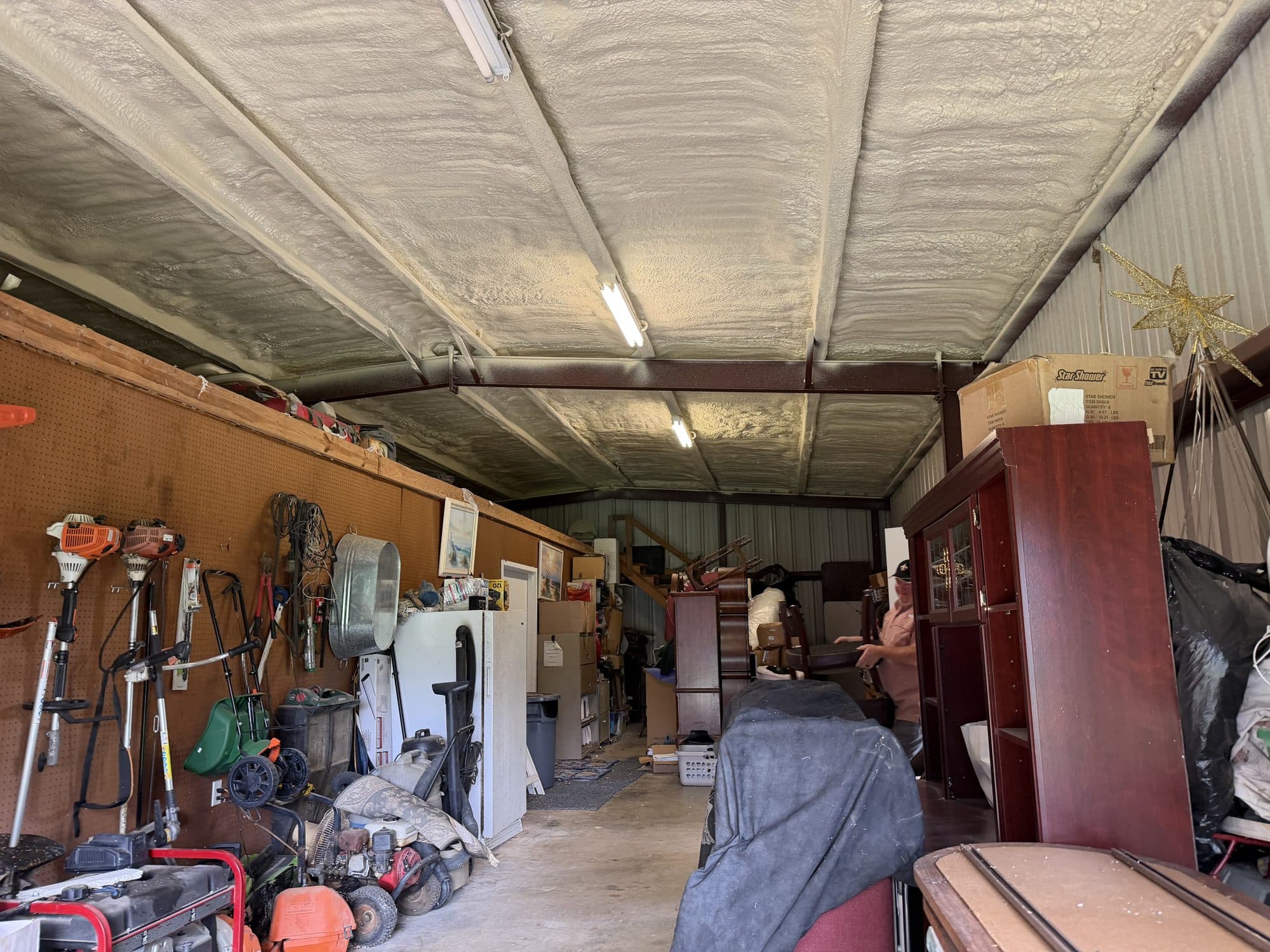 Cluttered garage with spray foam insulation, garden tools on a pegboard, and stored furniture.