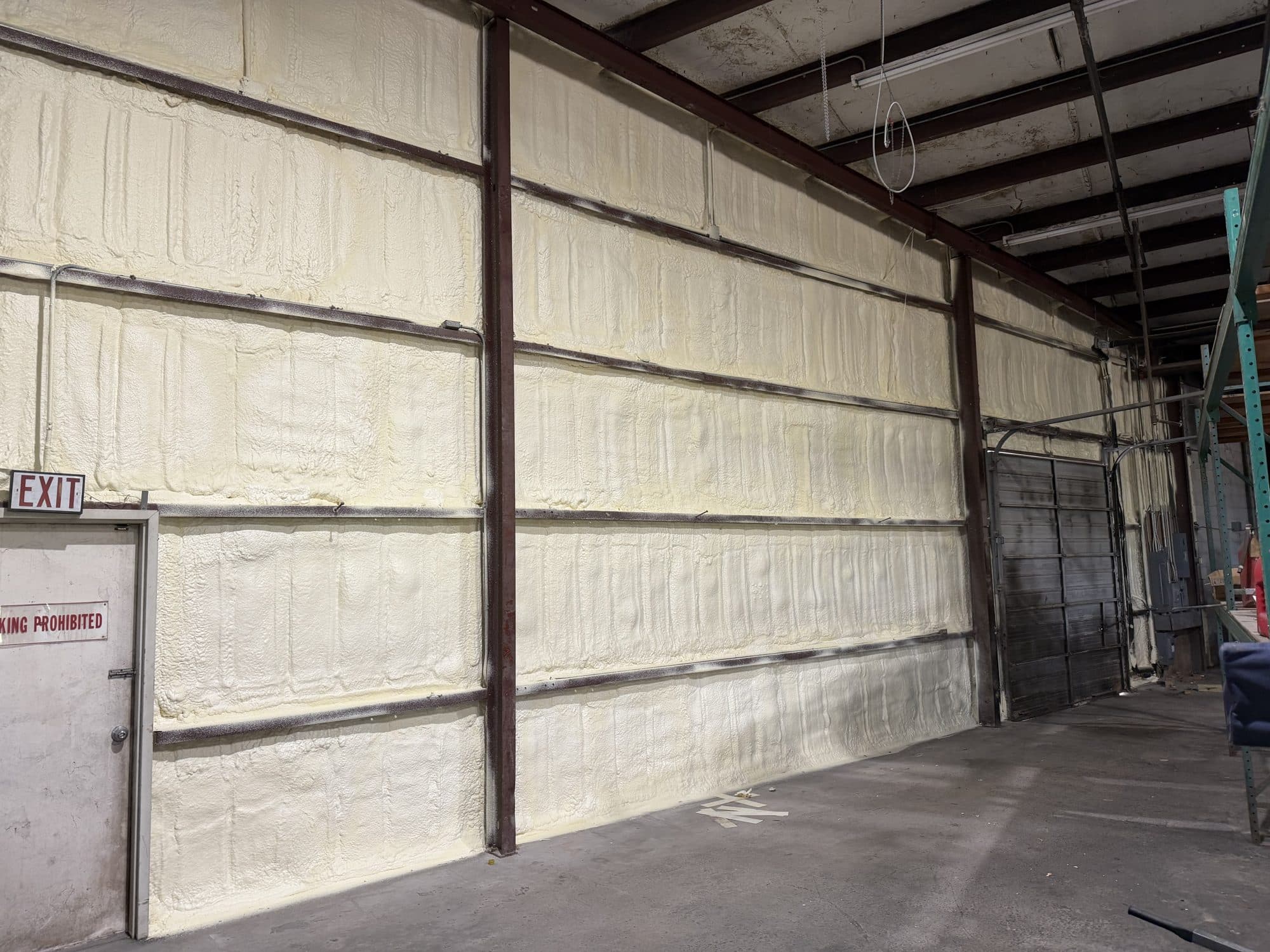 Interior warehouse wall covered in thick off-white spray foam insulation between metal structural beams.