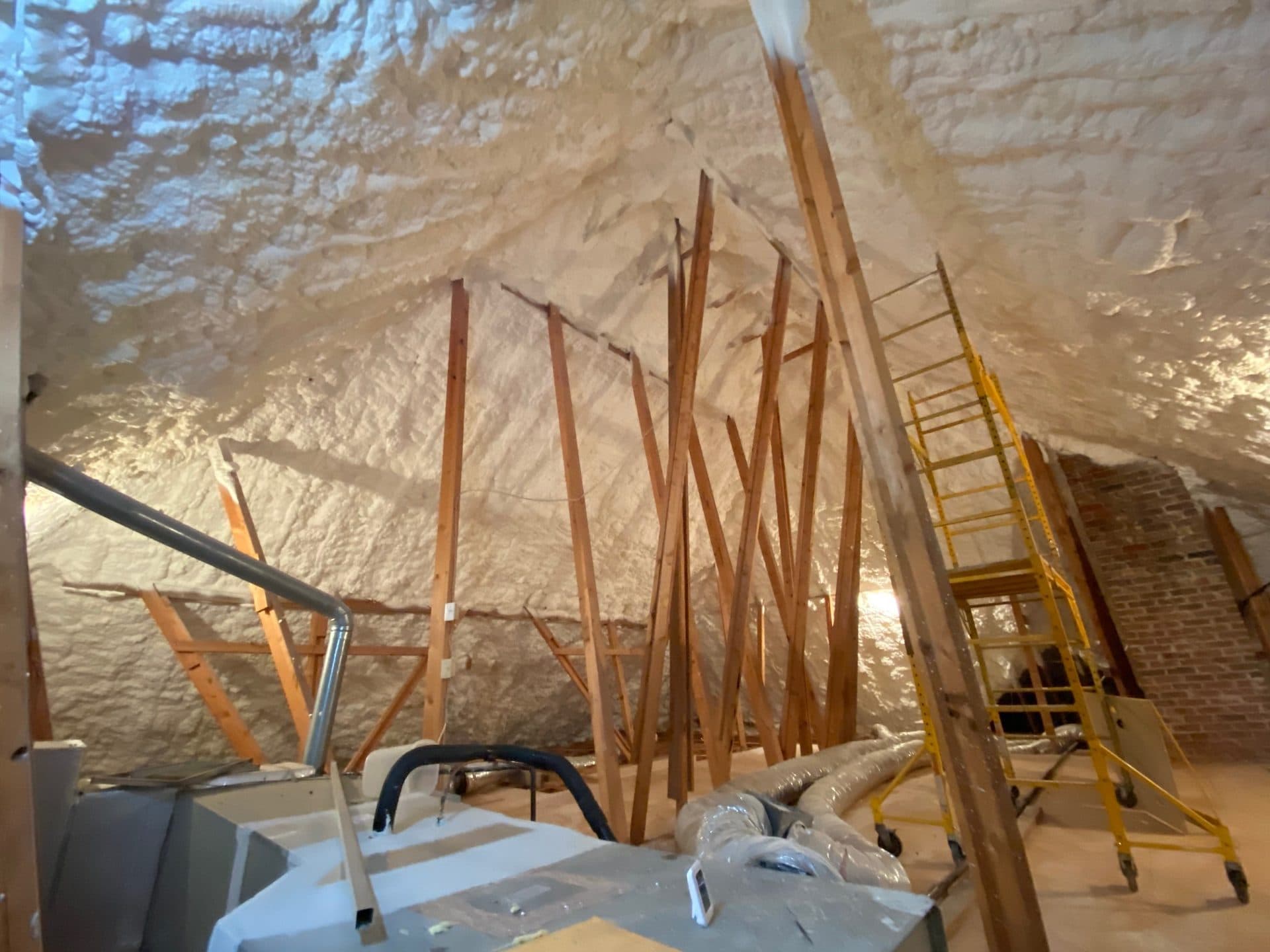 Attic space with white spray foam insulation, wooden support beams, and yellow rolling ladder.
