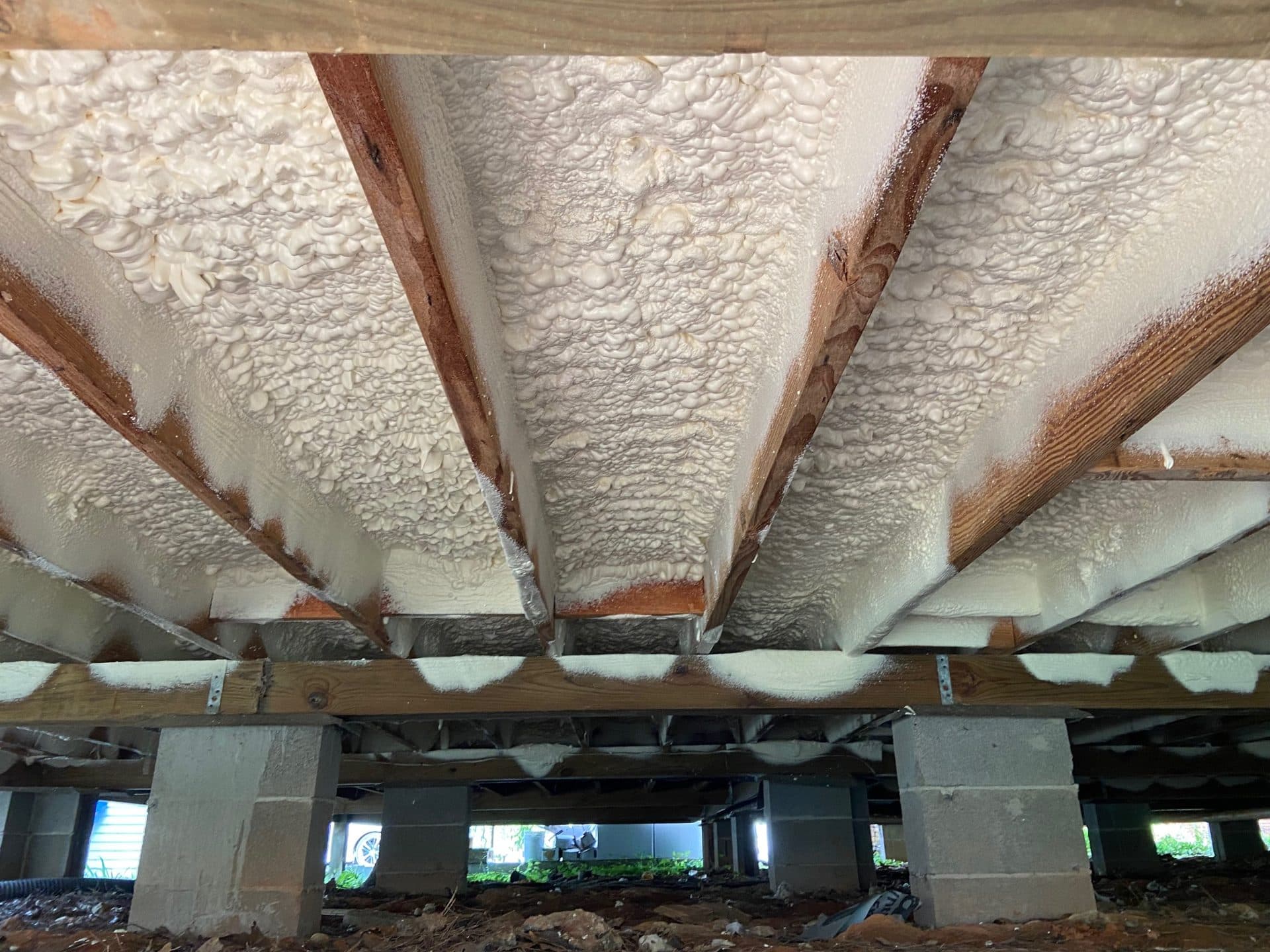 White spray foam insulation applied between wooden floor joists in a residential crawl space.