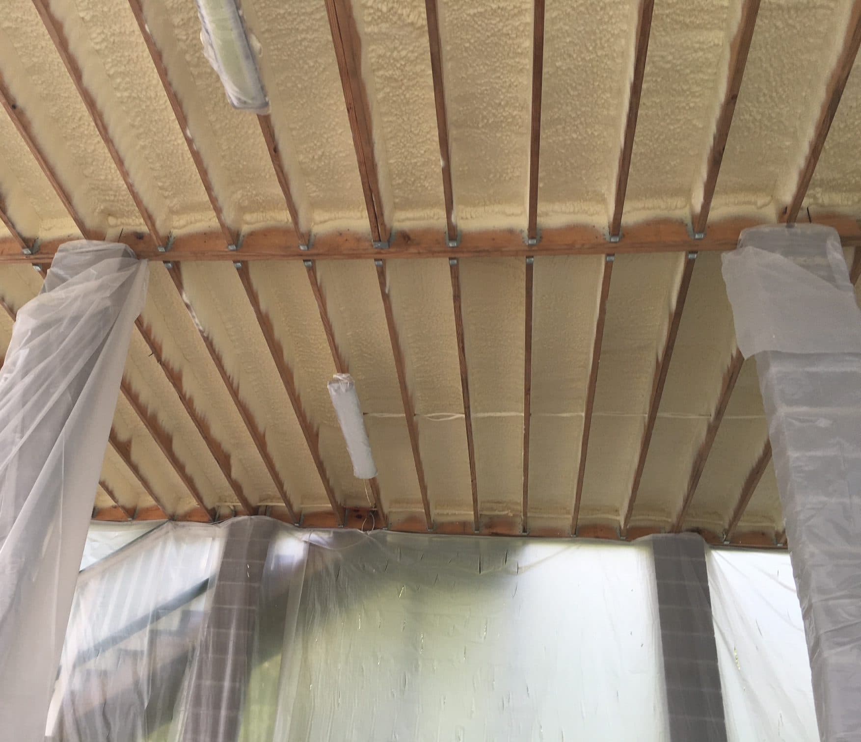Off-white spray foam insulation applied between wooden ceiling joists with protective plastic sheeting nearby.