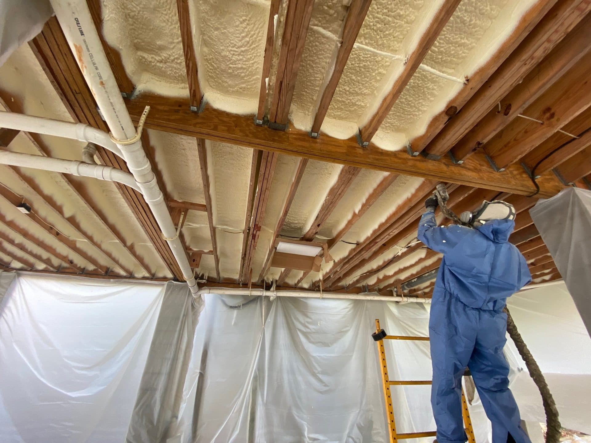 Worker in blue suit applying spray foam insulation between wooden ceiling joists in a crawlspace.