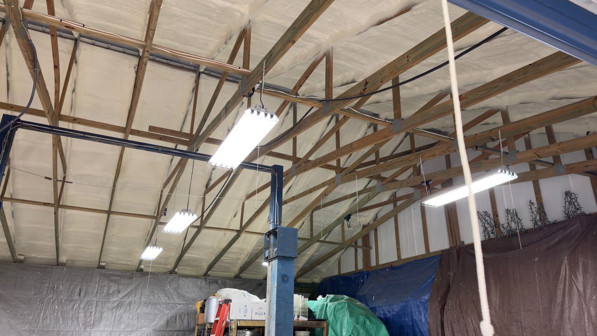 Bright LED shop lights hang from wooden trusses in a spray foam insulated workshop.