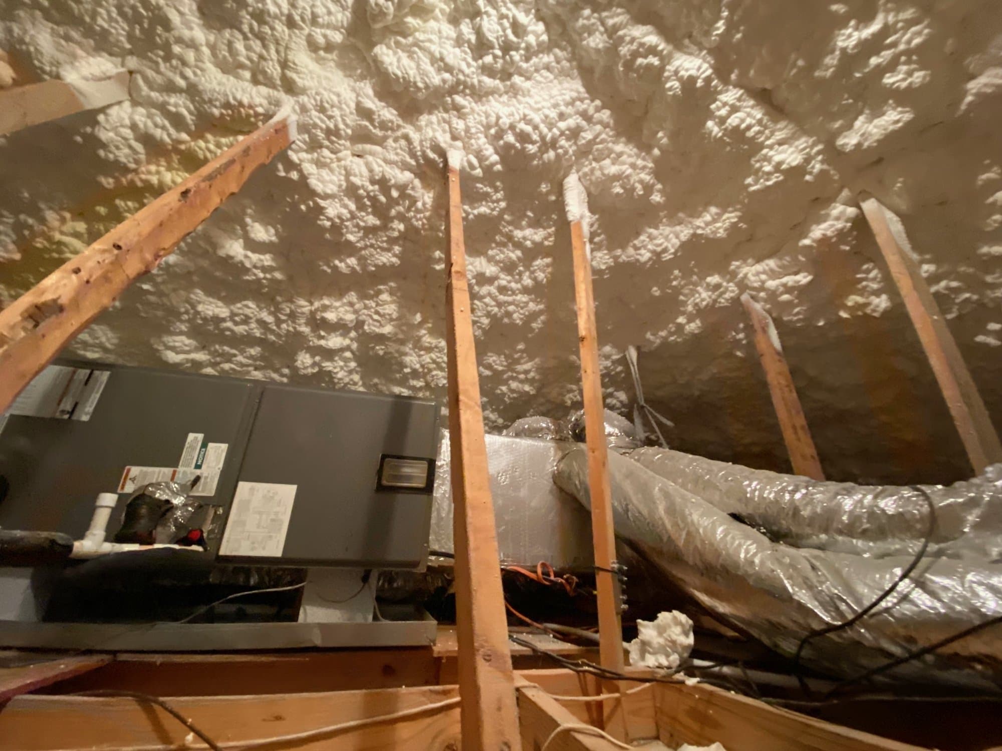 White spray foam insulation covers an attic roof deck above an HVAC unit and ductwork.