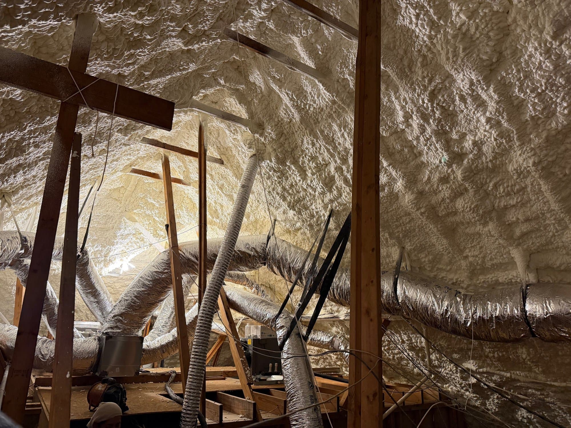 Attic roof covered in white spray foam insulation with silver flexible ducts and wooden framing.