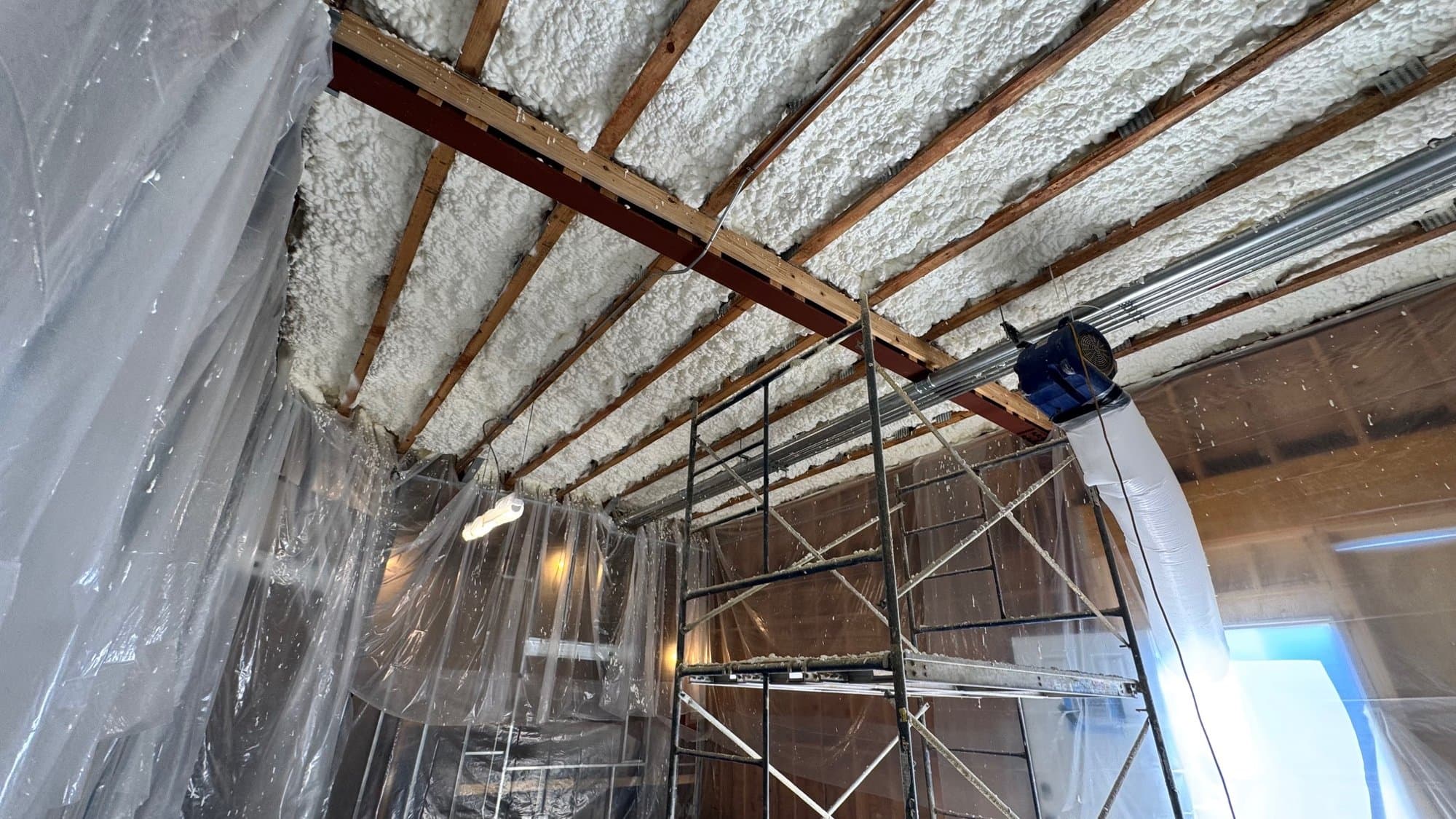 Spray foam insulation on ceiling rafters above scaffolding and plastic sheeting in a construction site.