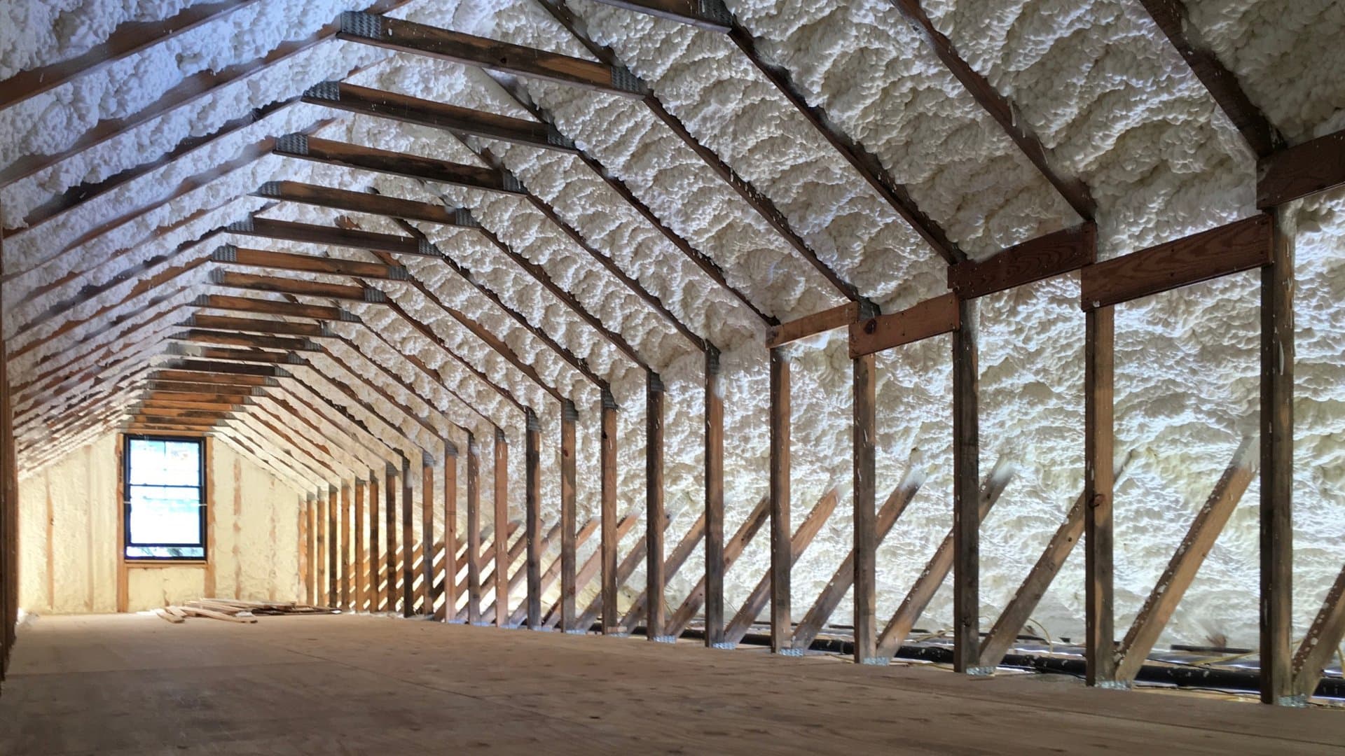 Attic interior with white spray foam insulation between wooden rafters and a small window.