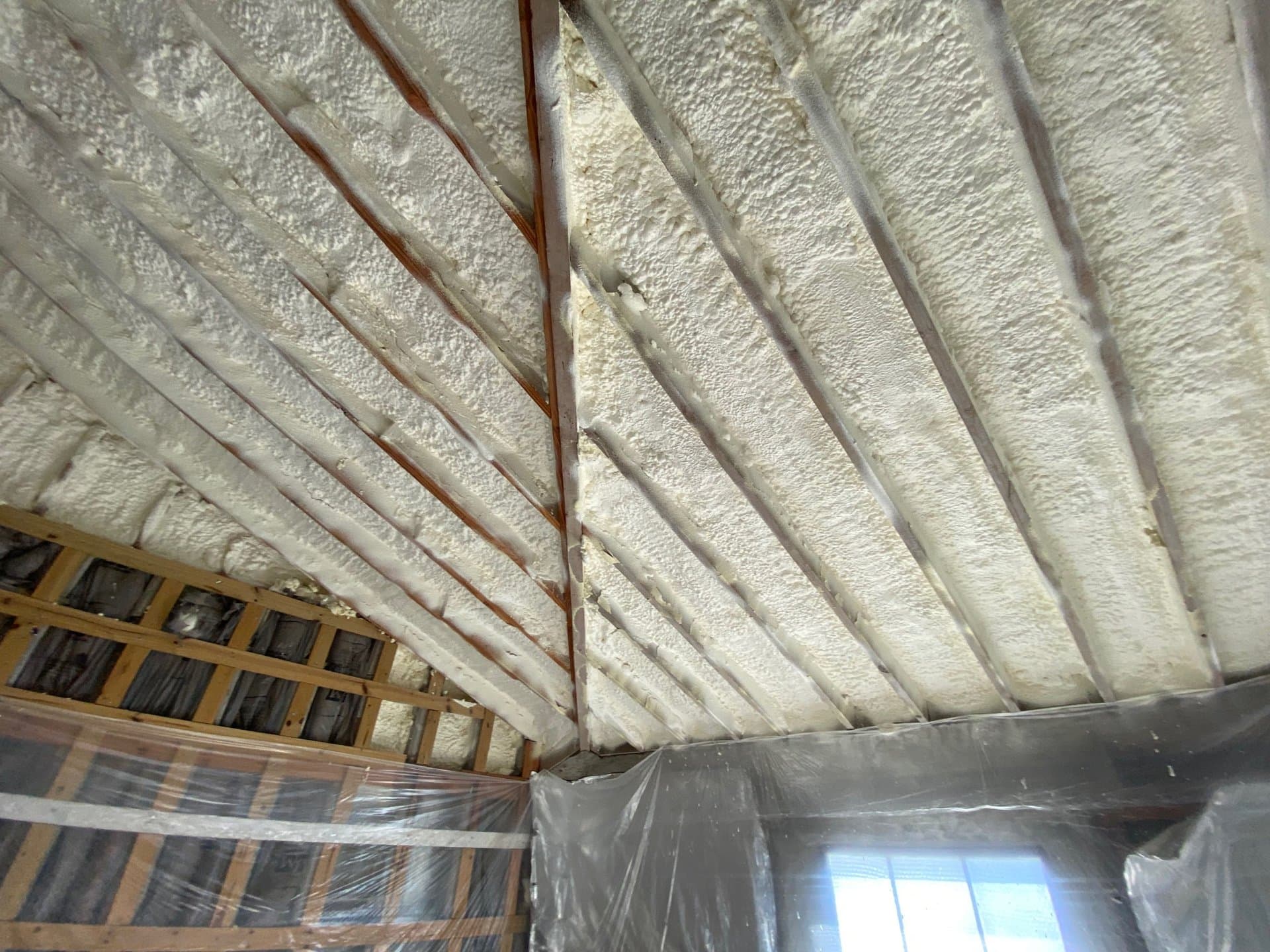 Thick white spray foam insulation fills the gaps between wooden rafters in a vaulted ceiling.