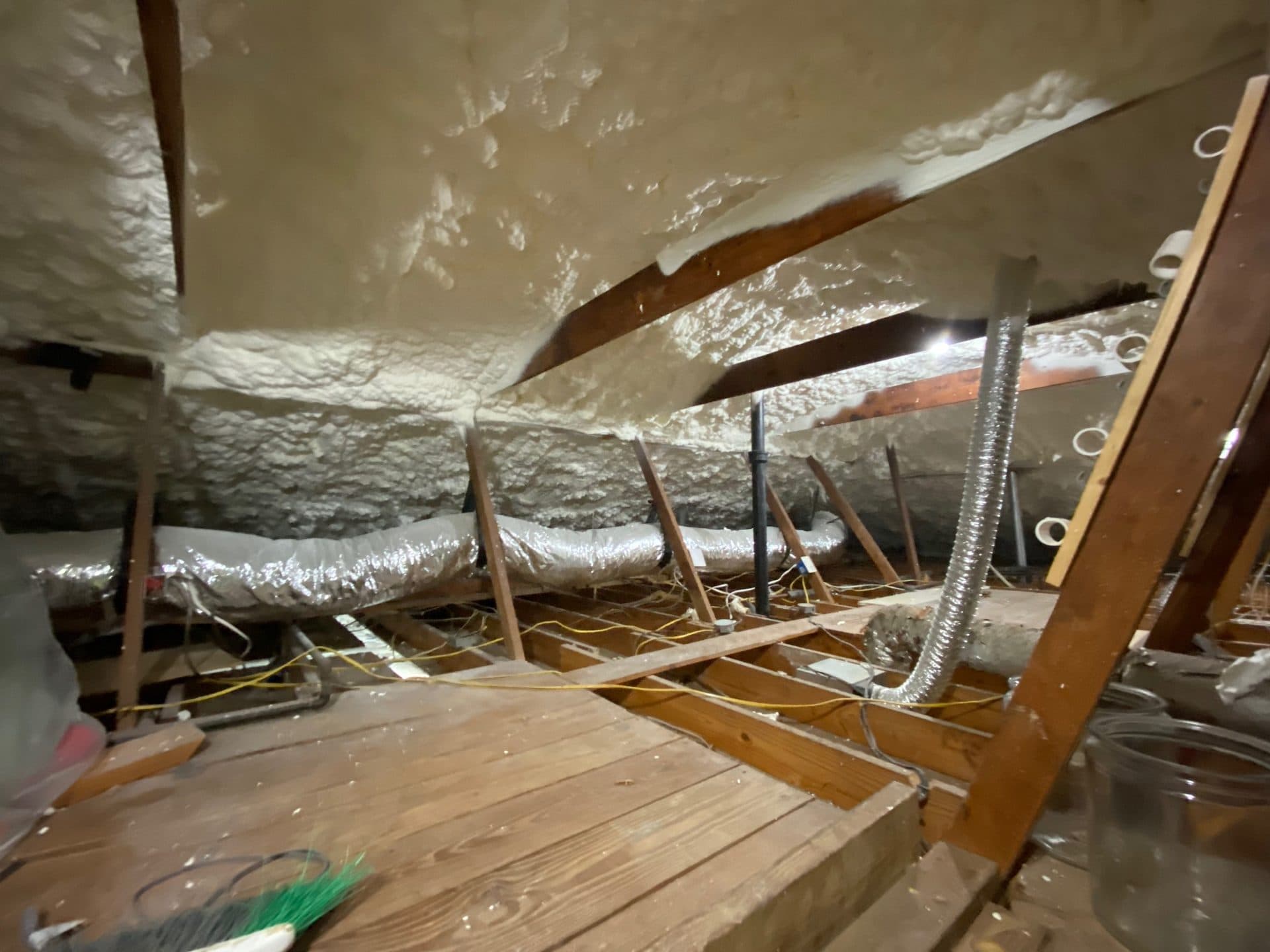 Attic interior featuring white spray foam insulation, wooden rafters, and flexible silver ventilation ducts.