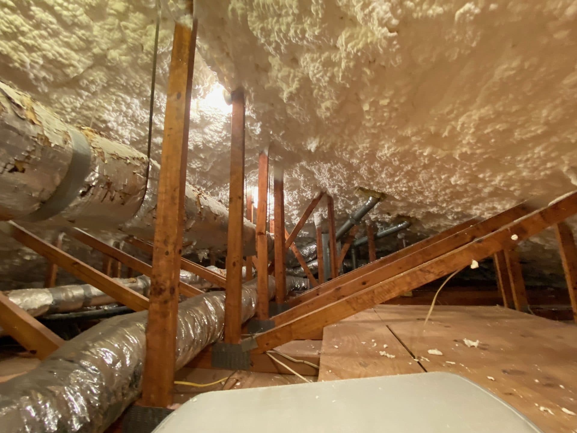 White spray foam insulation covers an attic ceiling above wooden rafters and silver HVAC ducts.