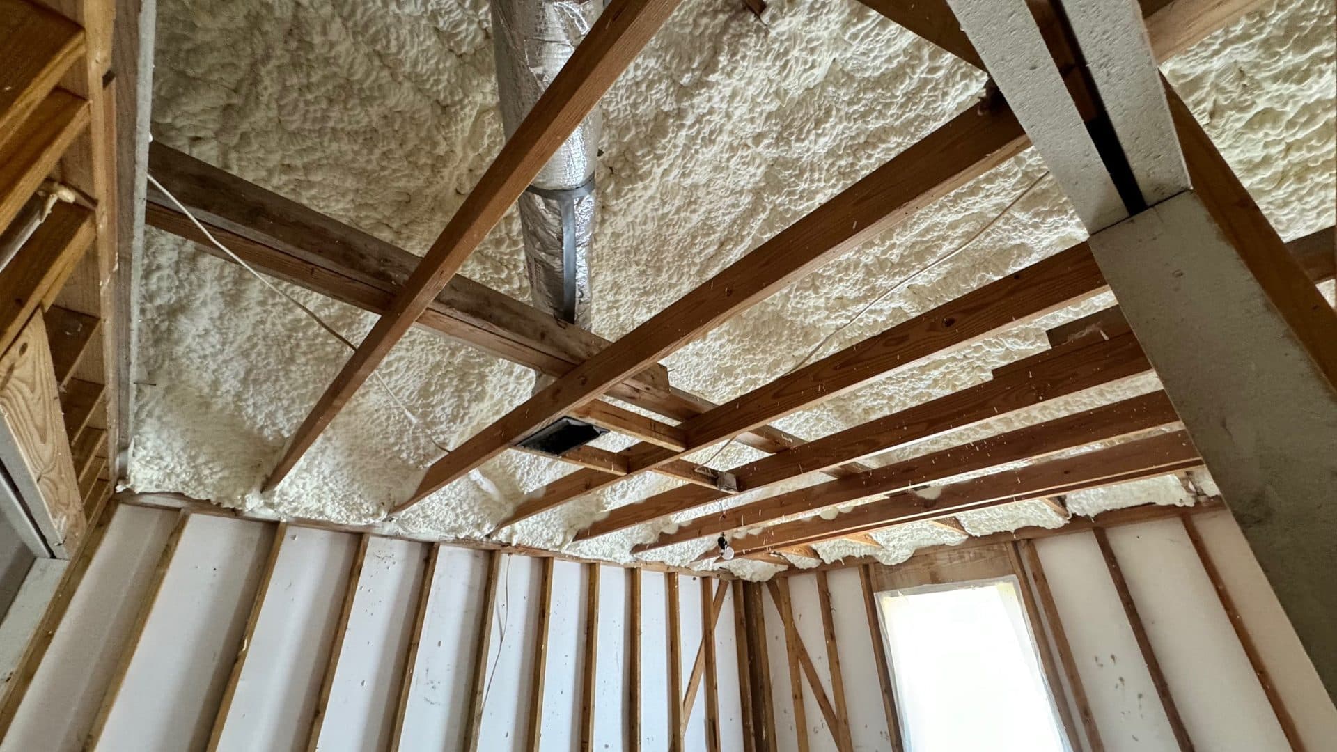 Thick white spray foam insulation applied between wooden ceiling joists in an unfinished attic.
