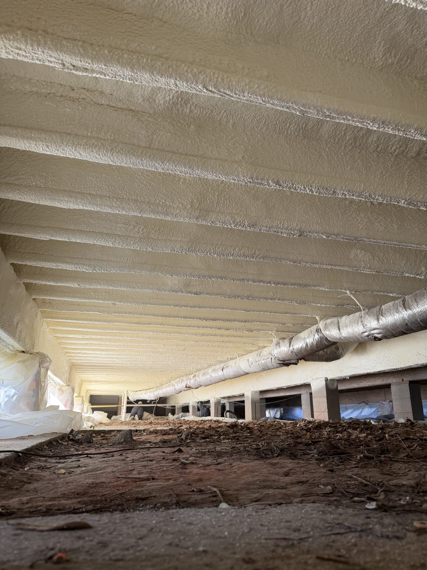 White spray foam insulation covers the ceiling of a crawl space with insulated ductwork.