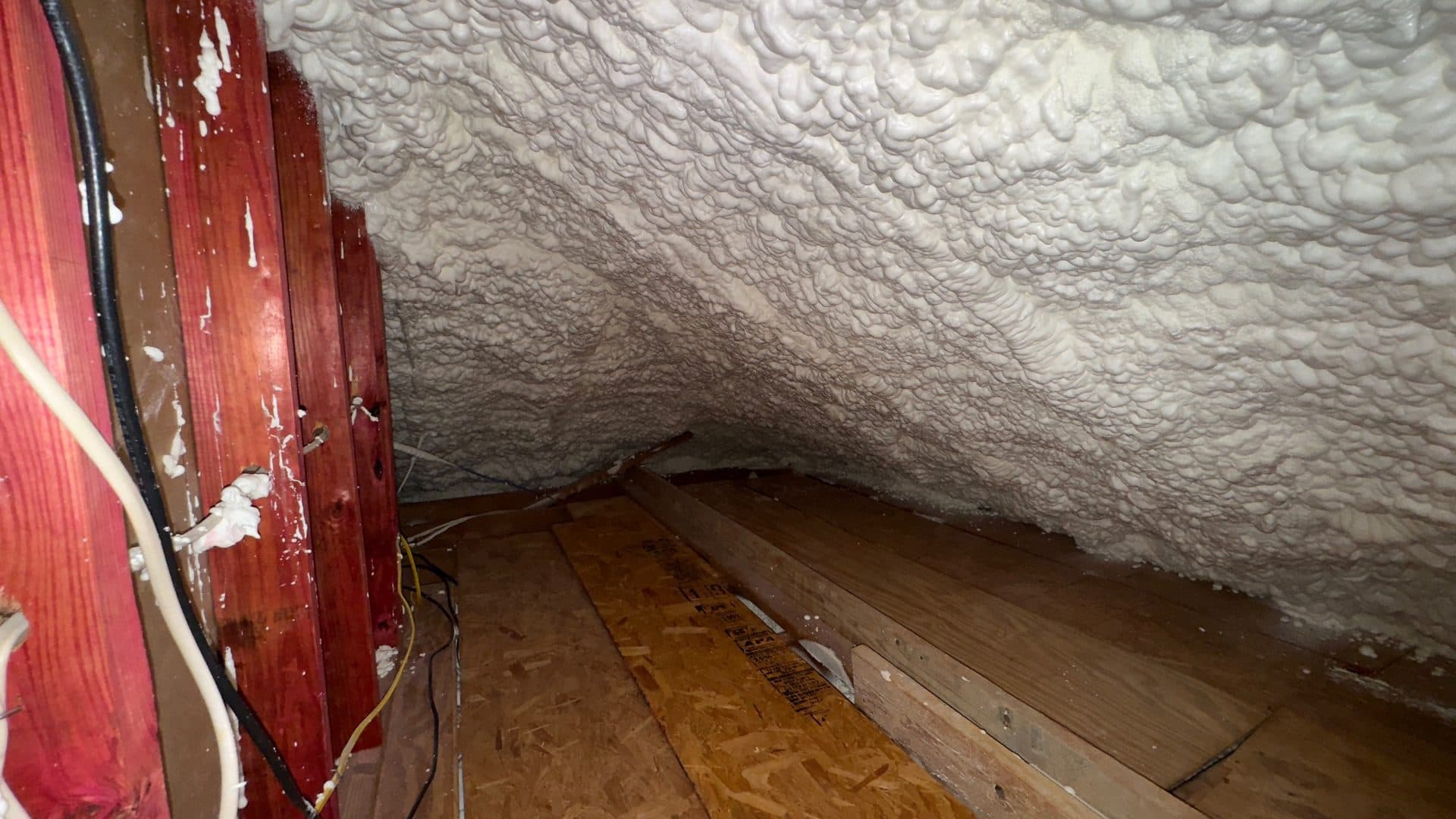 White spray foam insulation covers the ceiling of an attic with wooden floorboards and studs.