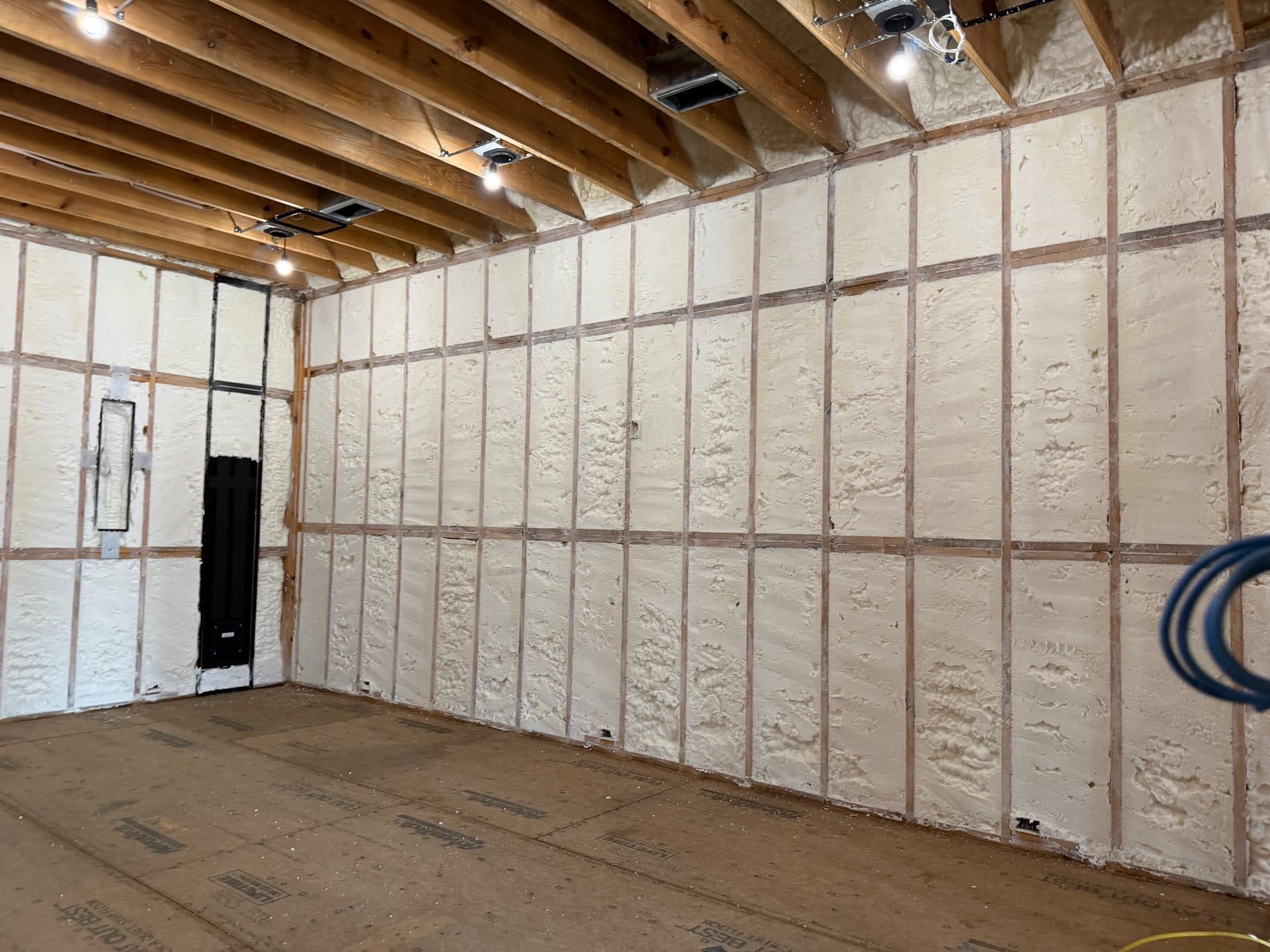 Interior room under construction with spray foam insulation between wooden wall studs and exposed ceiling joists.
