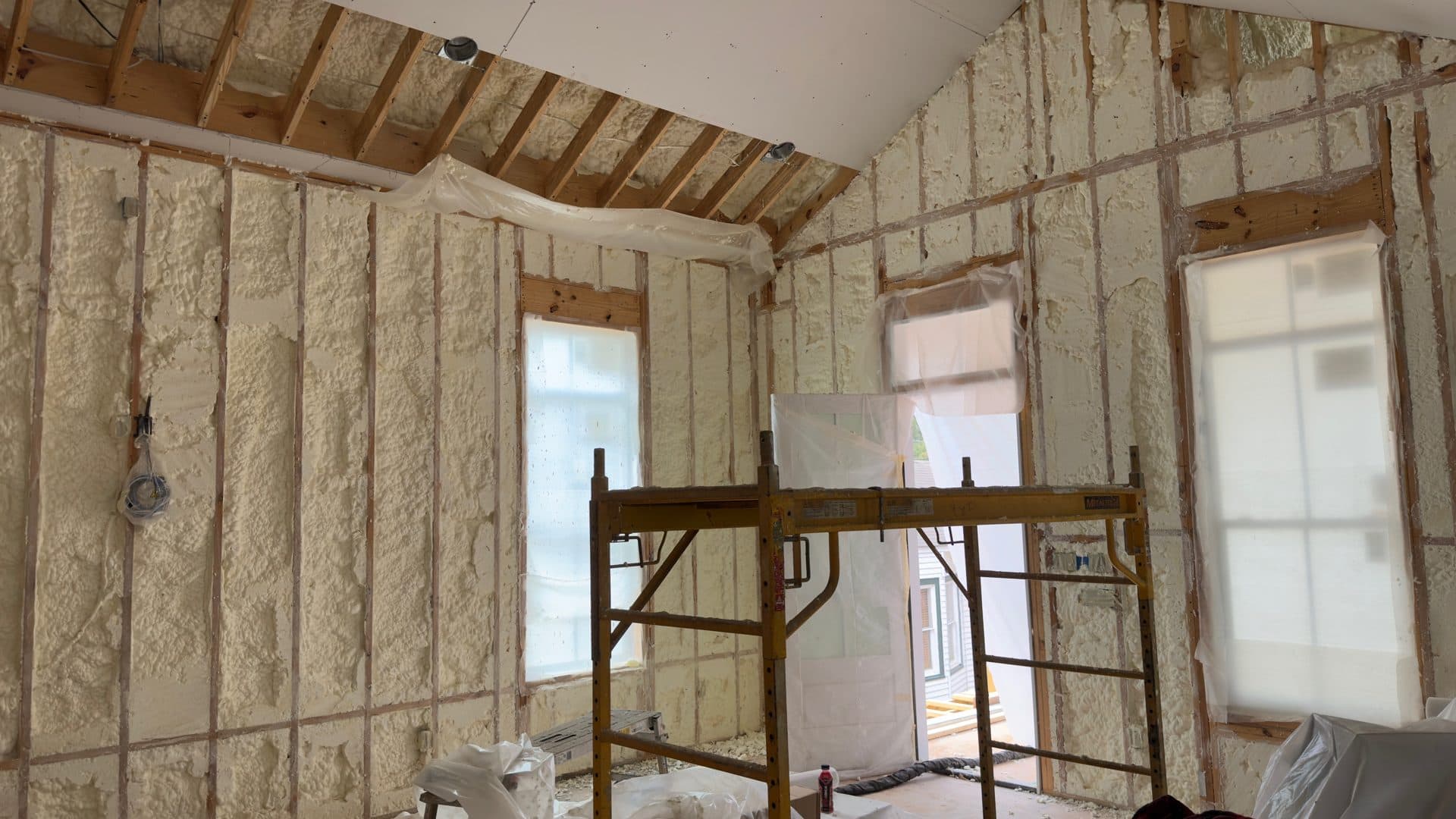 Interior room under construction with spray foam insulation between wooden studs and yellow scaffolding.