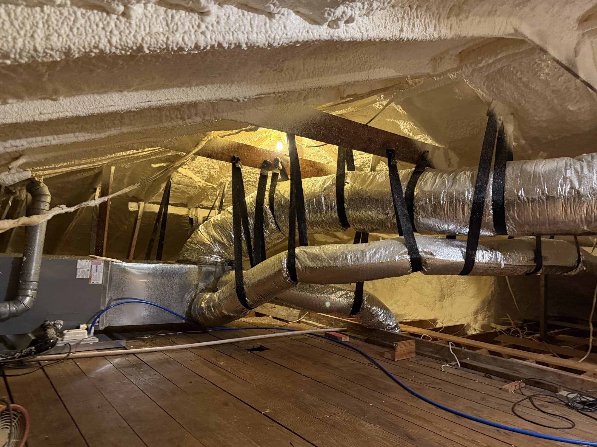 Spray foam insulated attic with HVAC unit and flexible ducts suspended by black straps.