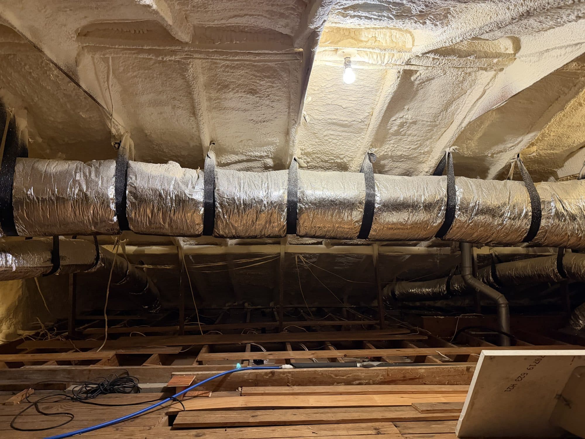 Attic with white spray foam insulation on the ceiling and silver flexible air conditioning ducts.