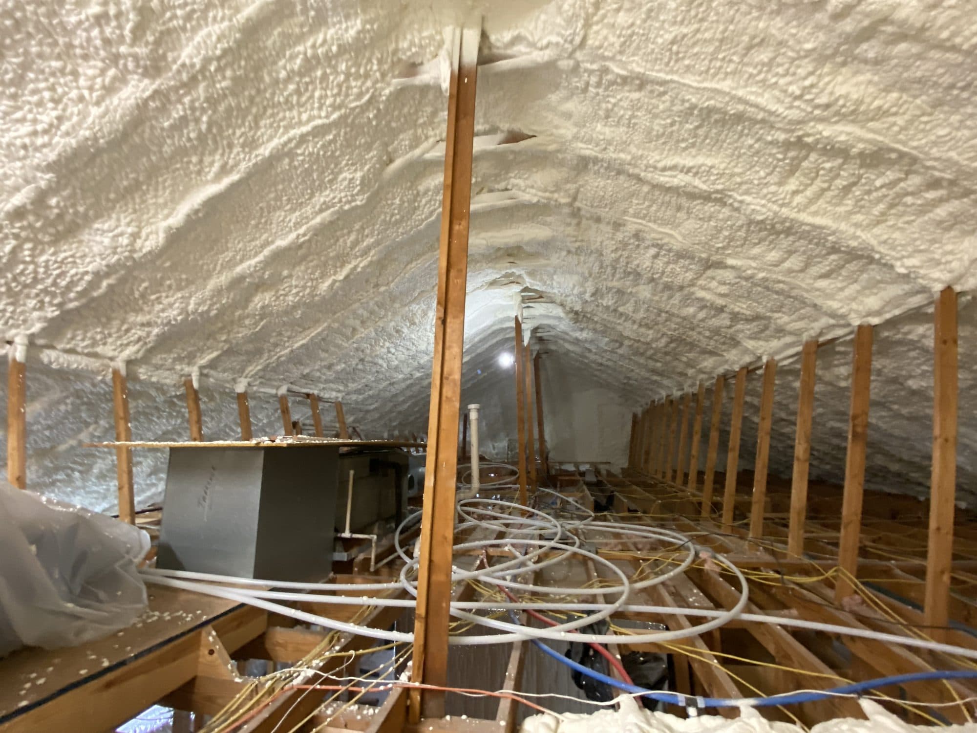 White spray foam insulation lines an attic roof above wooden framing and floor utility pipes.