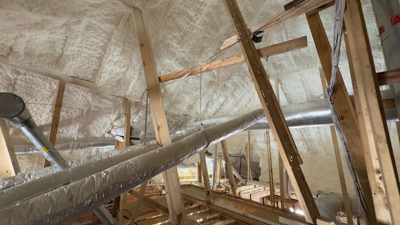 White spray foam insulation applied to an attic roof deck with wooden framing and ductwork.