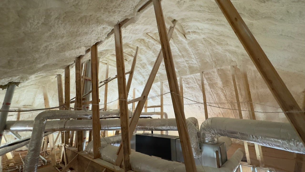 Thick white spray foam insulation covers an attic's roof and walls around wooden support beams.