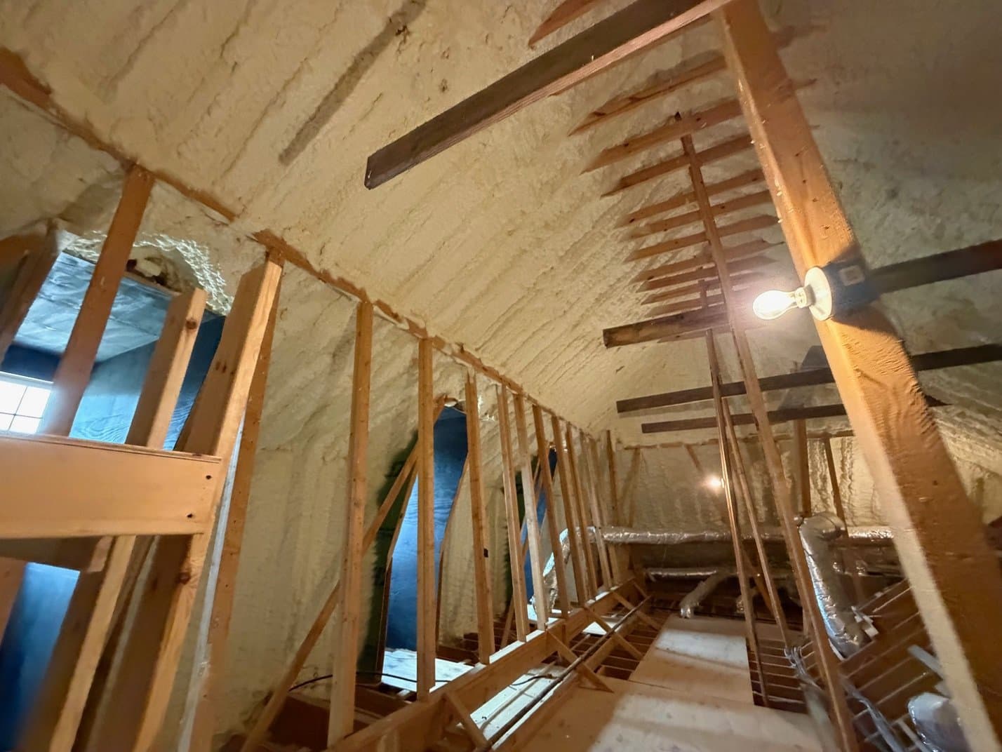 Spray foam insulation covers the ceiling and walls of a wooden-framed attic space under construction.