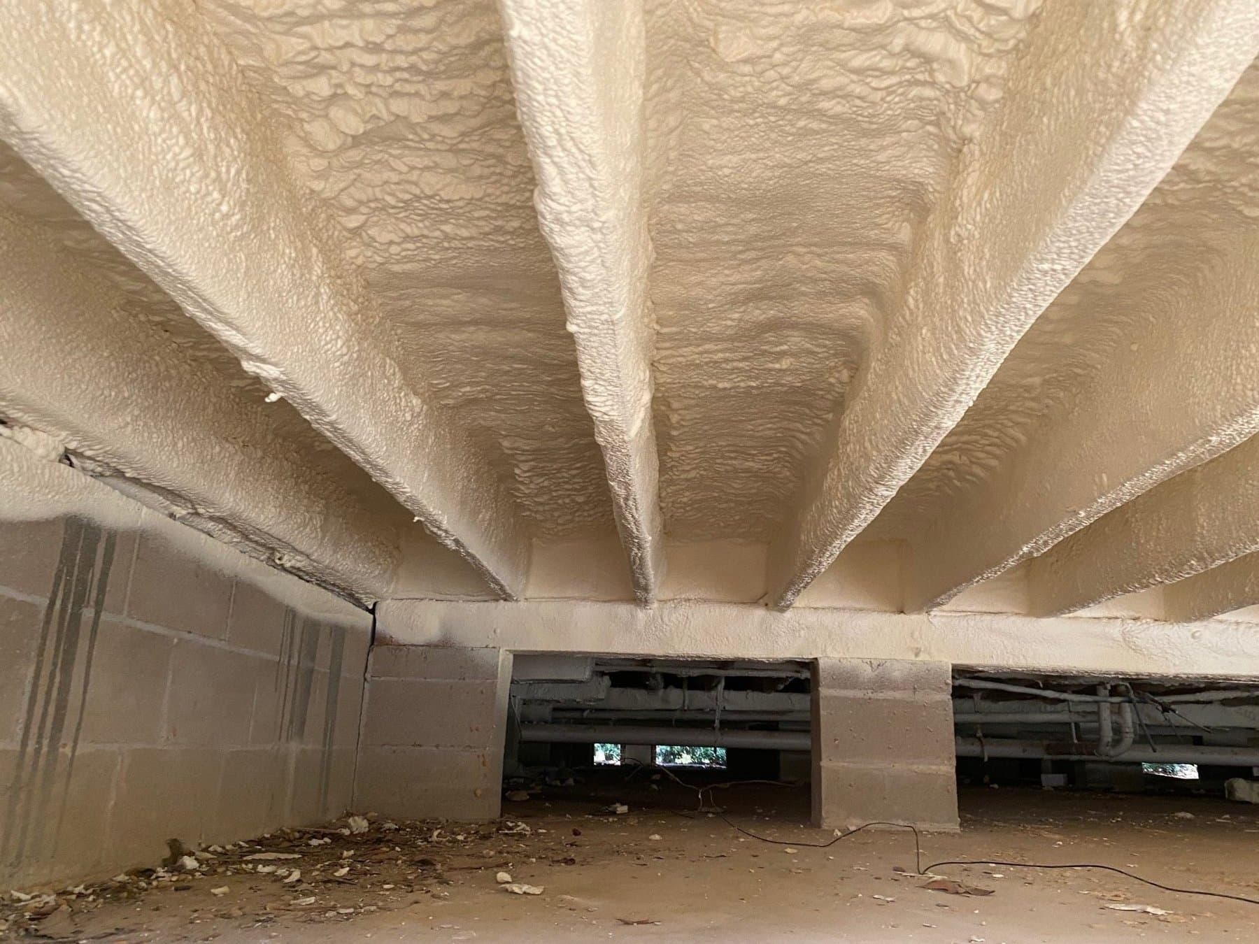 Off-white spray foam insulation applied to floor joists and subflooring in a crawl space.
