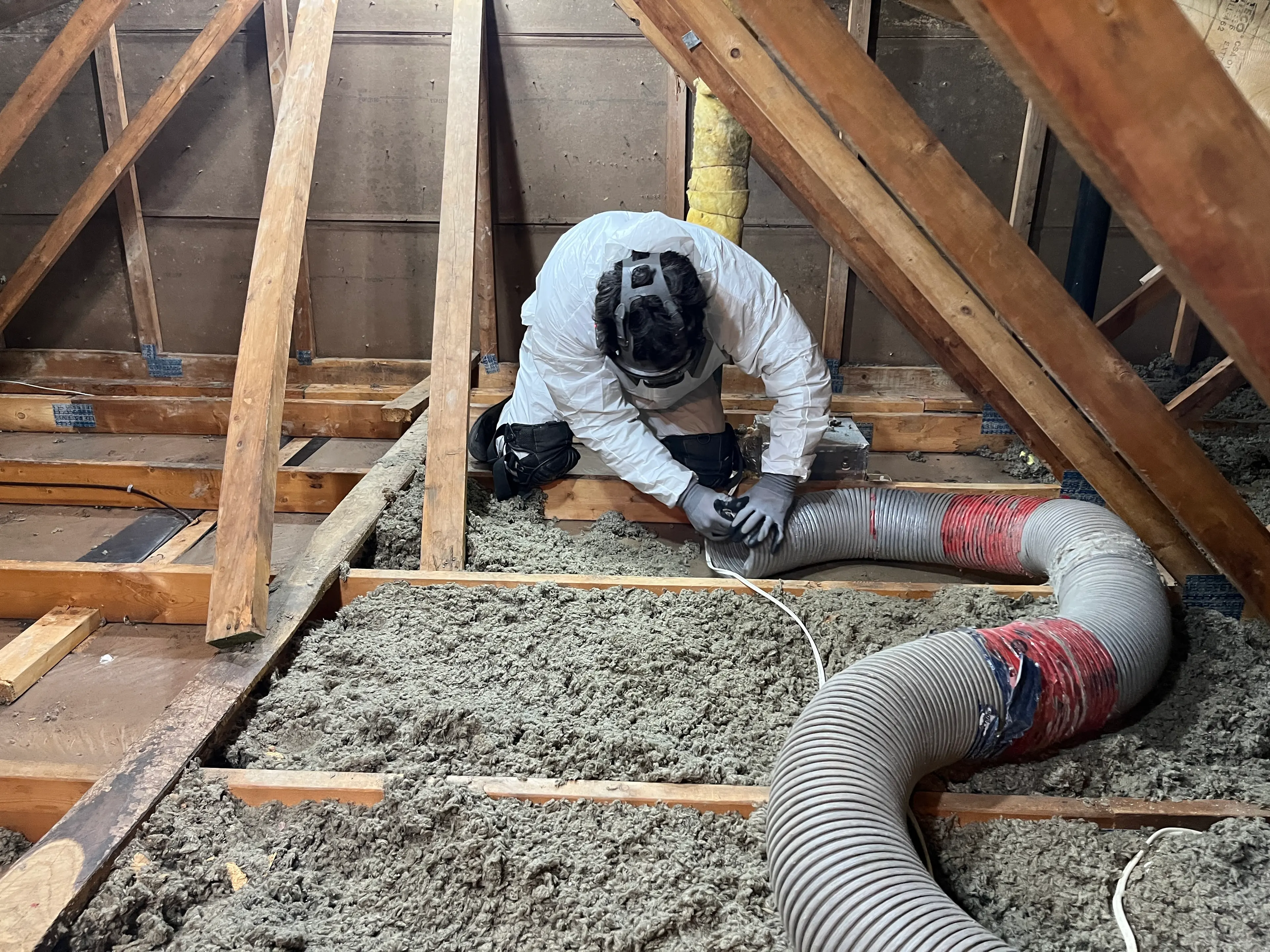 Worker in protective gear installs loose-fill insulation in an attic using a large hose.