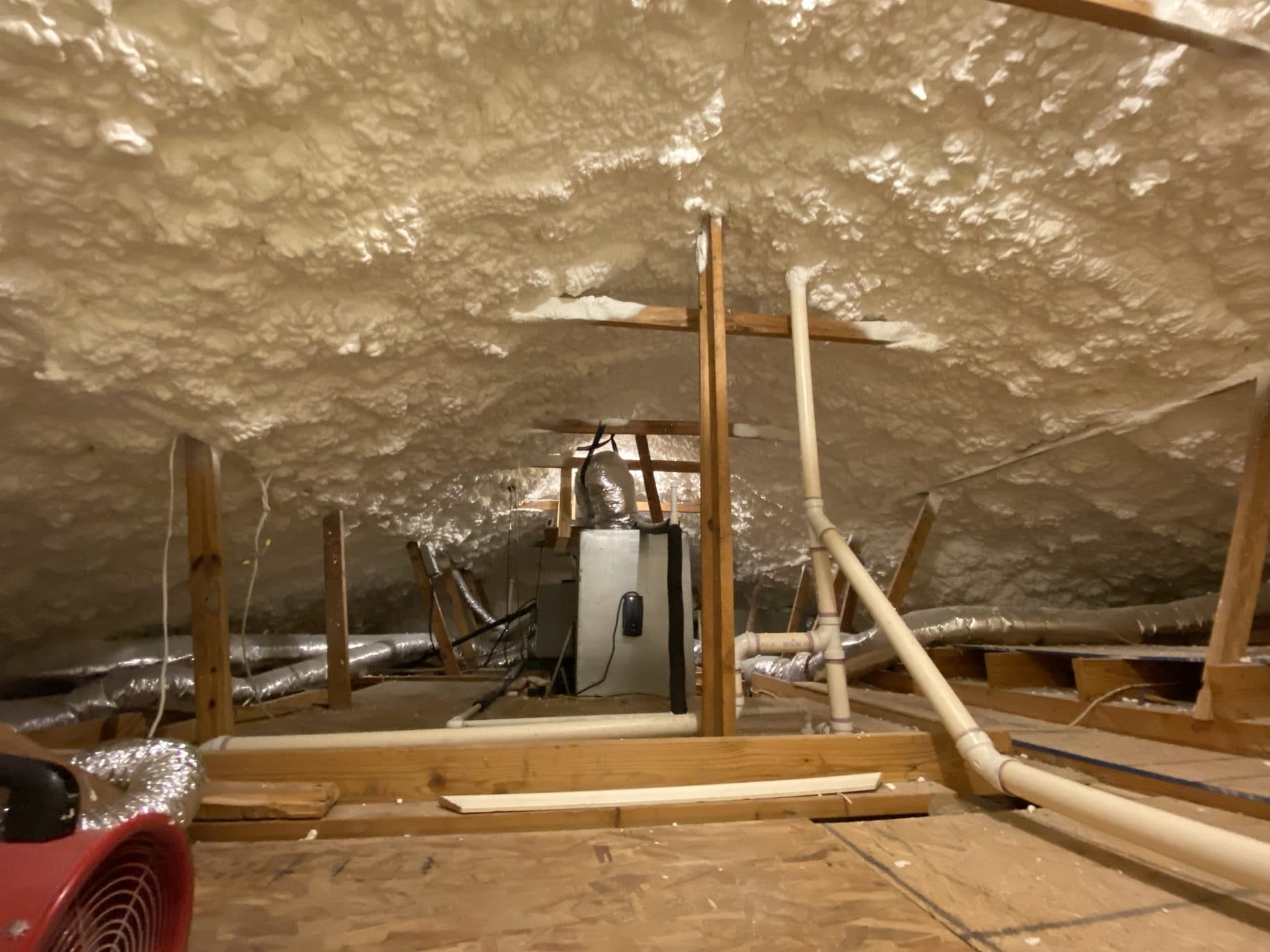 Attic interior with white spray foam insulation, wooden beams, an HVAC unit, and utility pipes.