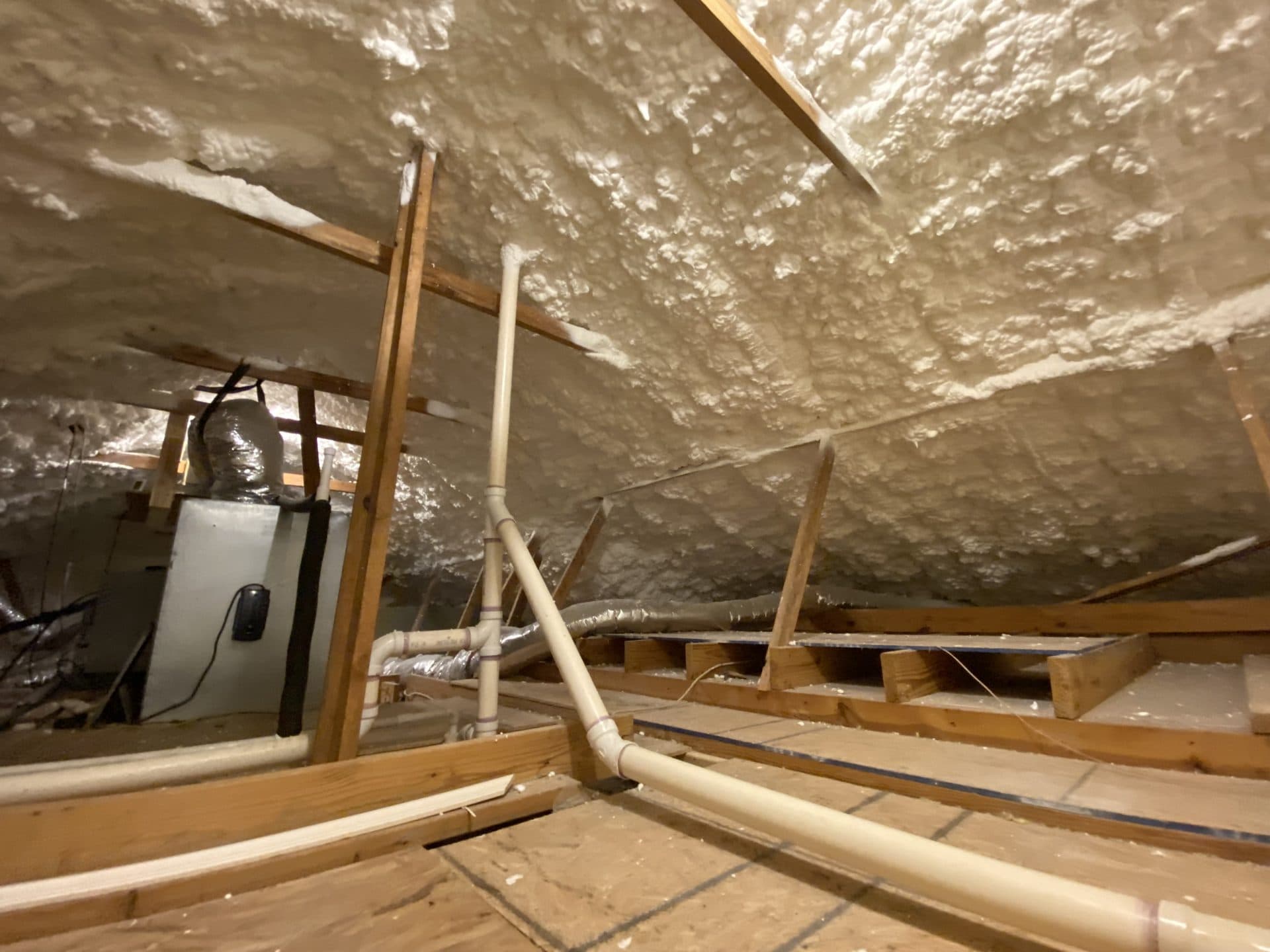 Attic space with white spray foam insulation on the roof, wooden rafters, and PVC pipes.