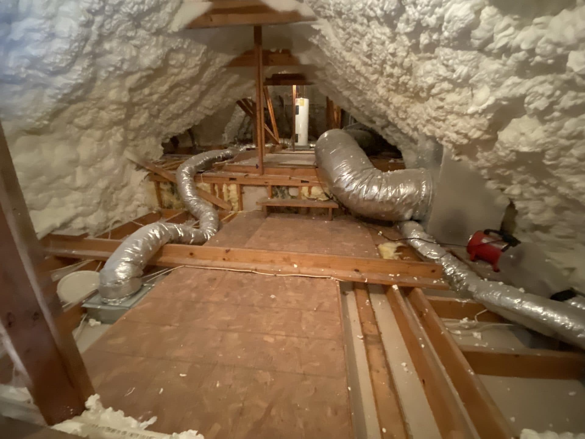Attic with white spray foam insulation and silver flexible HVAC ducts on wooden floorboards.