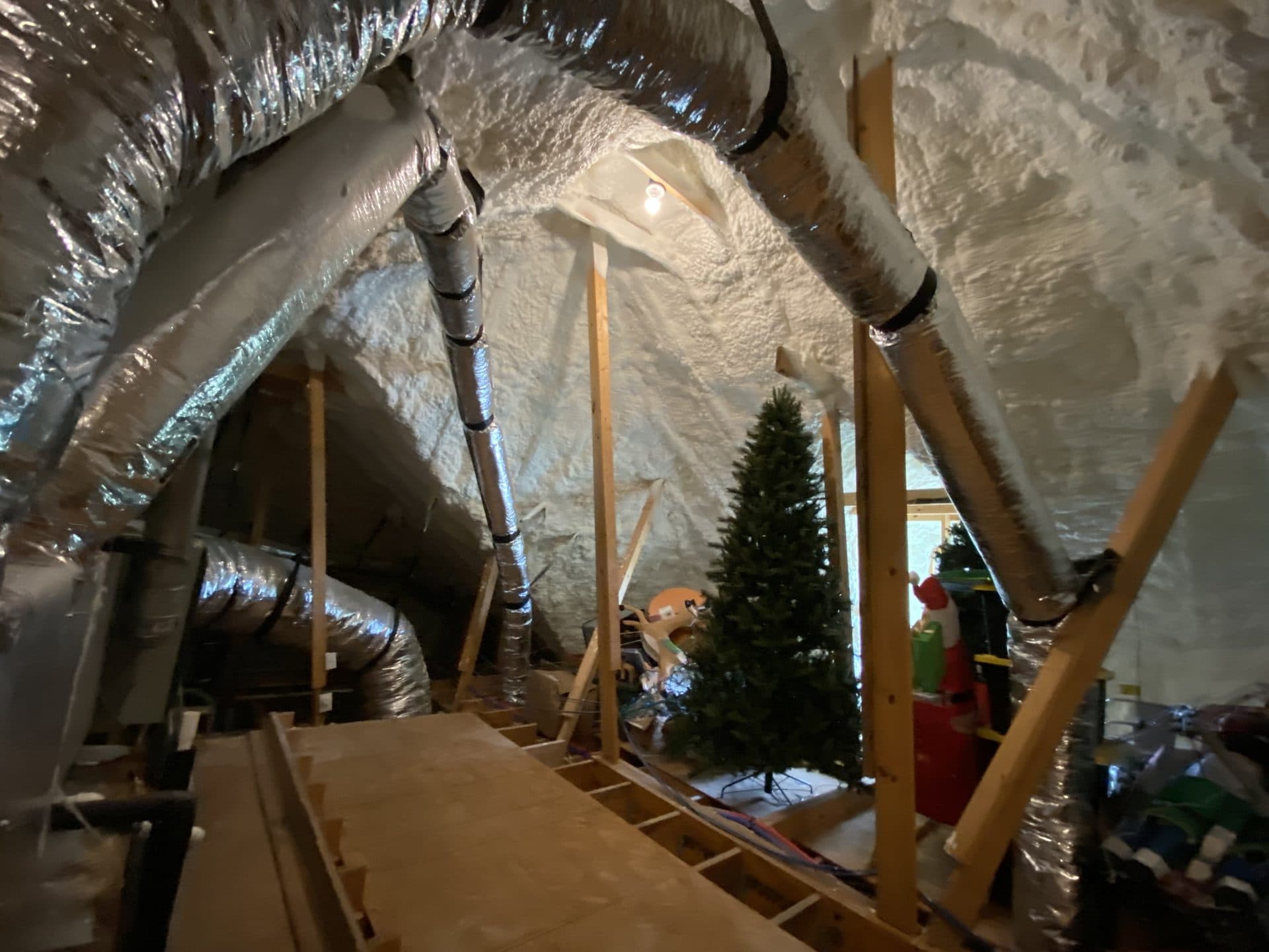 Attic interior featuring white spray foam insulation, silver ductwork, and a stored Christmas tree.