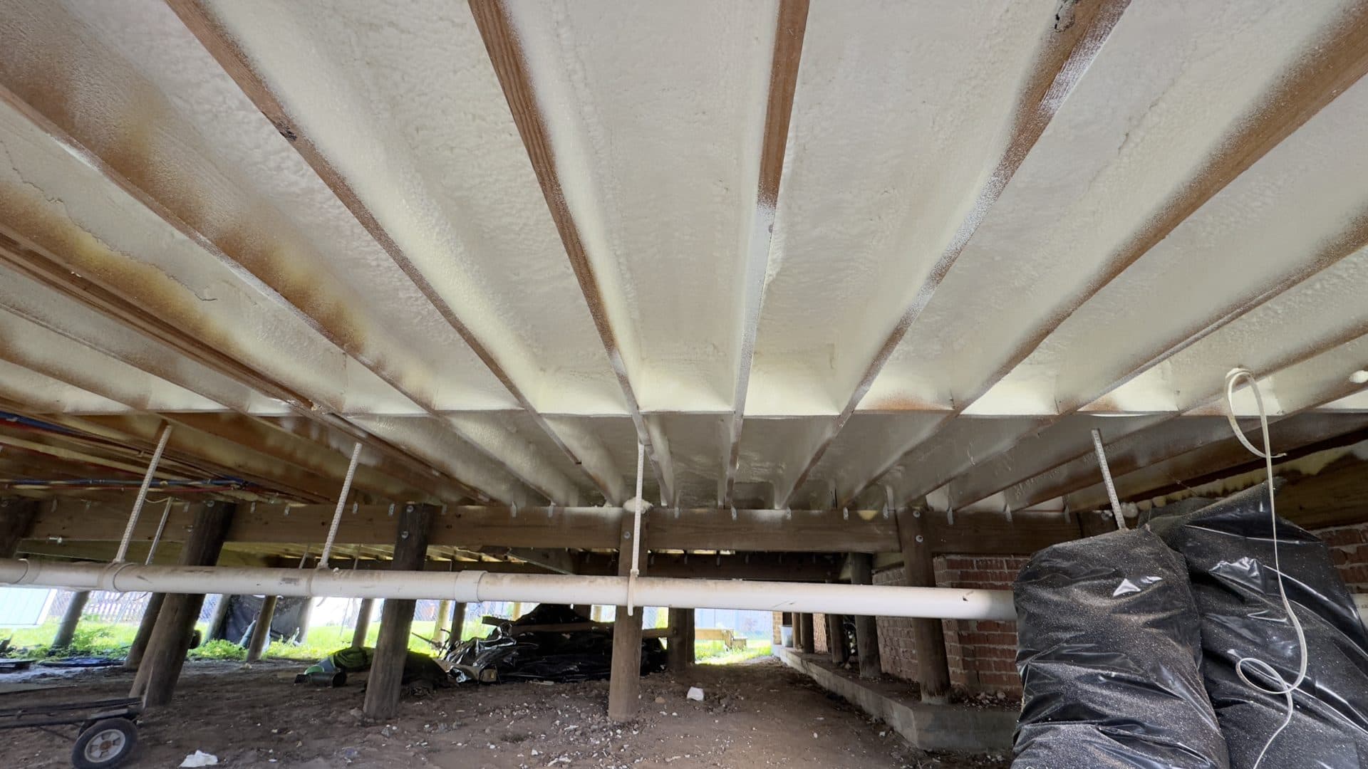 White spray foam insulation applied between wooden floor joists in a crawl space with pipes.