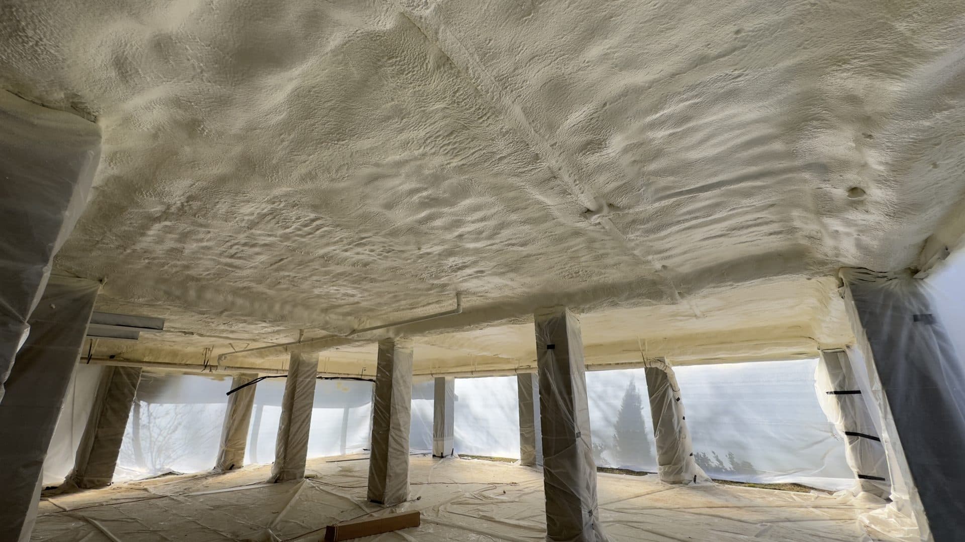 White spray foam insulation covers the ceiling of a crawlspace with plastic-wrapped support pillars.