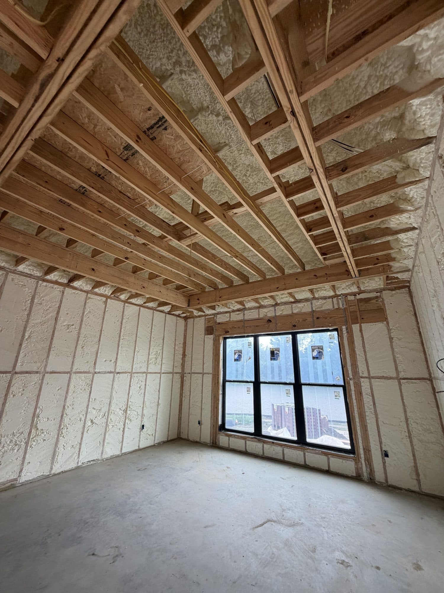 Interior room under construction with wood framing, spray foam insulation, and a large black window.