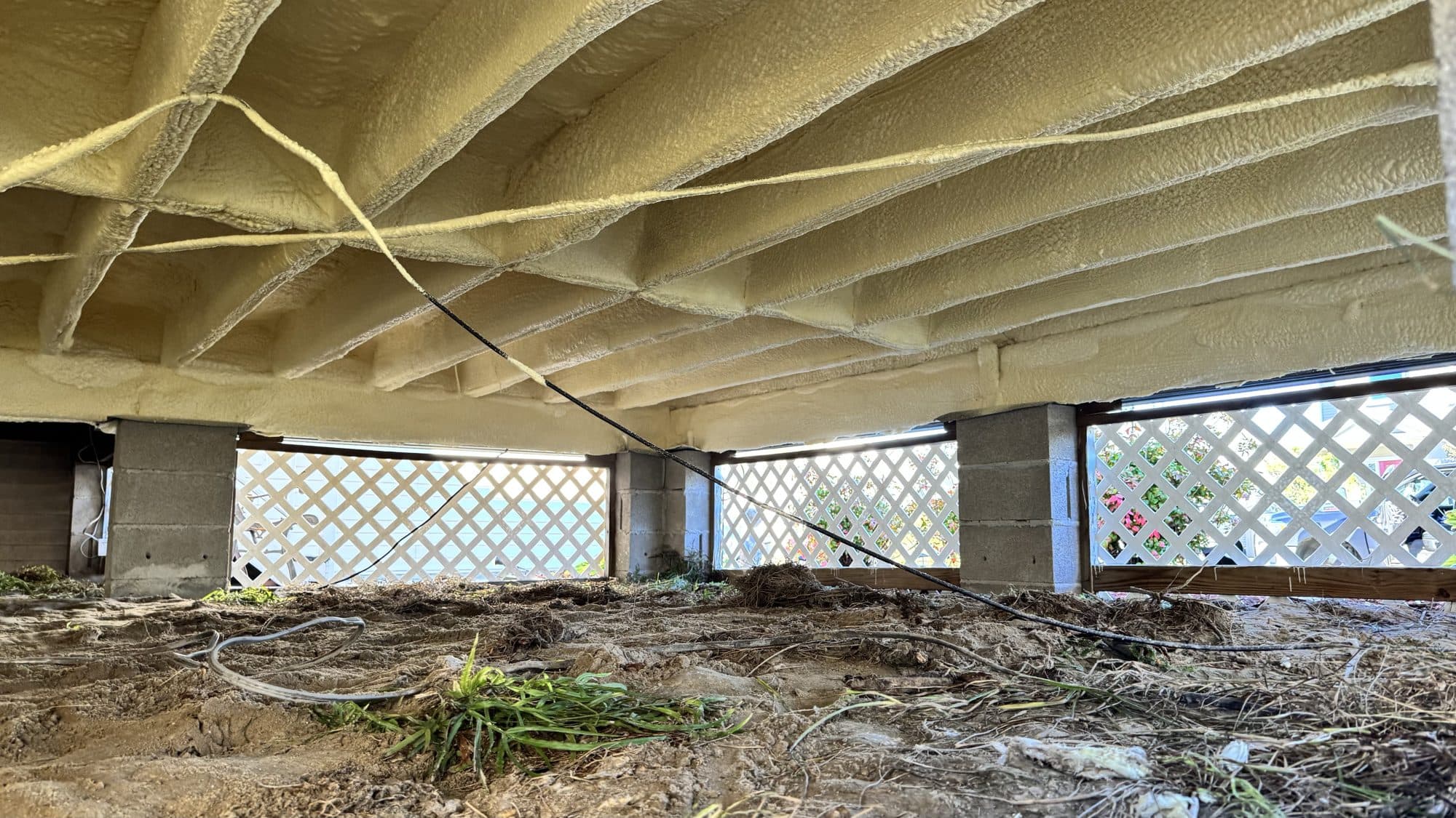 Yellow spray foam insulation on floor joists in a crawl space with dirt floor.